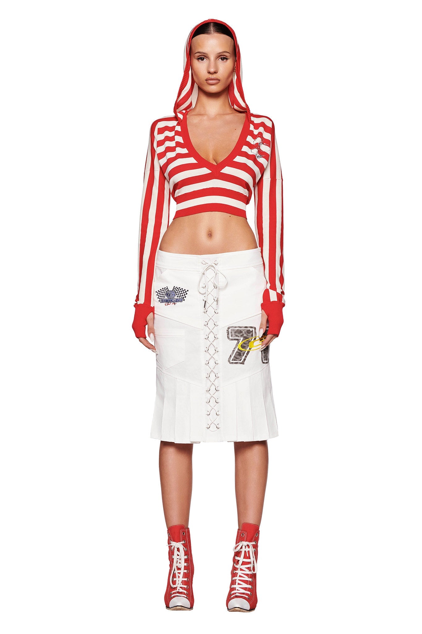 A woman wearing the SUZI SWEATER - RED : STRIPE and a white lace-up skirt from I.AM.GIA stands facing the camera. She has red fingerless gloves and matching high-top sneakers. She strikes a confident pose against a plain white background.