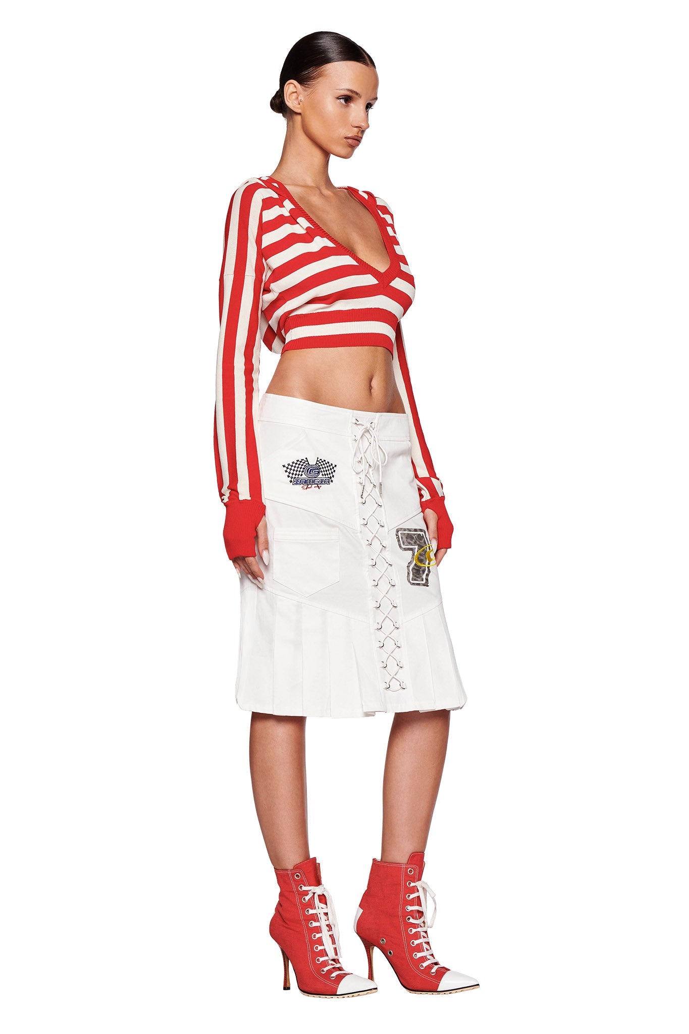 A woman stands against a white background, wearing the I.AM.GIA SUZI SWEATER in red and white stripes, paired with a white knee-length skirt featuring lace-up and patch details. She completes her outfit with red heeled ankle boots adorned with white laces, and her dark hair is styled sleekly back.