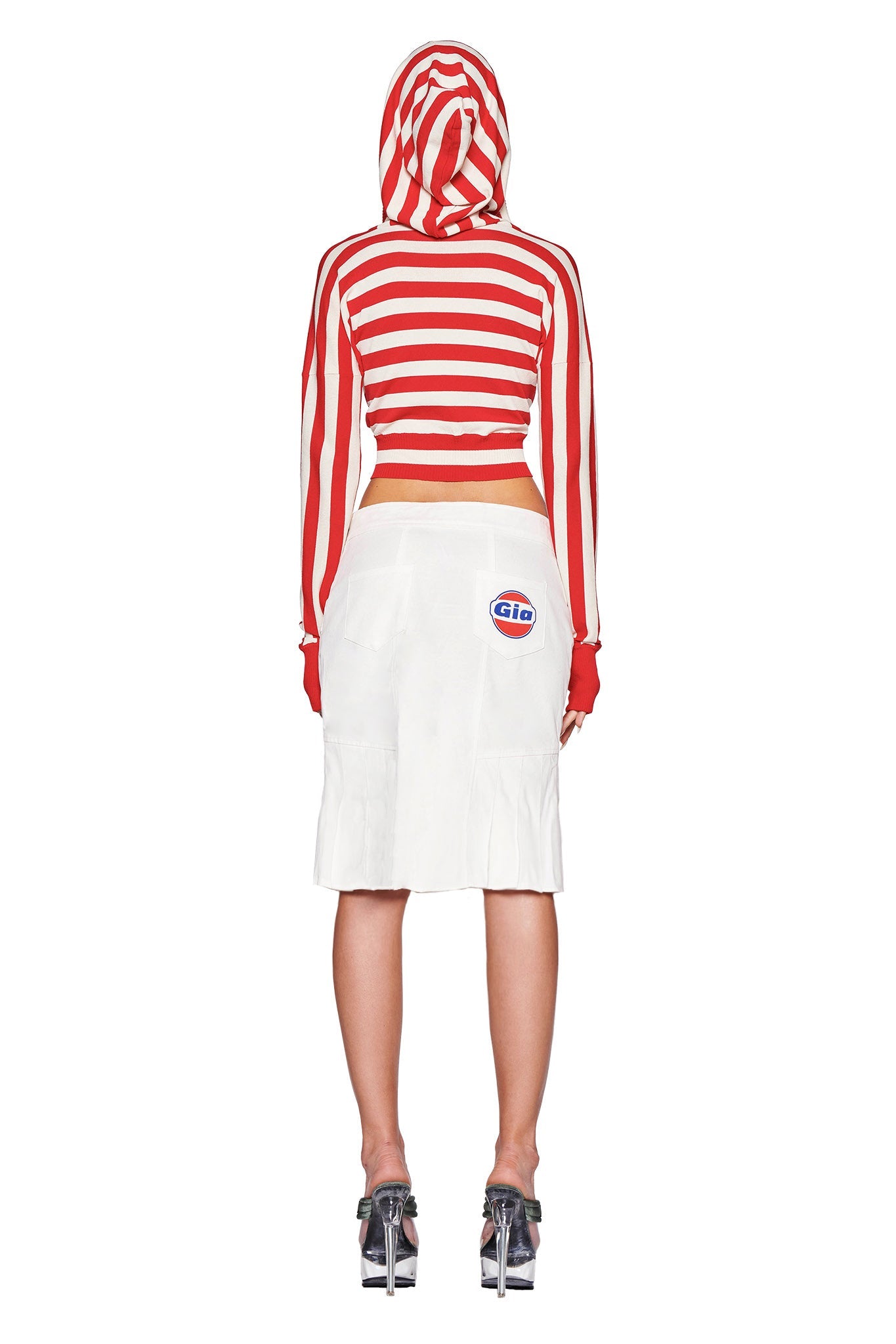 A person is standing with their back to the camera, dressed in the SUZI SWEATER - RED : STRIPE and a white skirt featuring a "CIAO" logo on the pocket. They are wearing high-heeled, transparent sandals from I.AM.GIA. The backdrop is plain white.