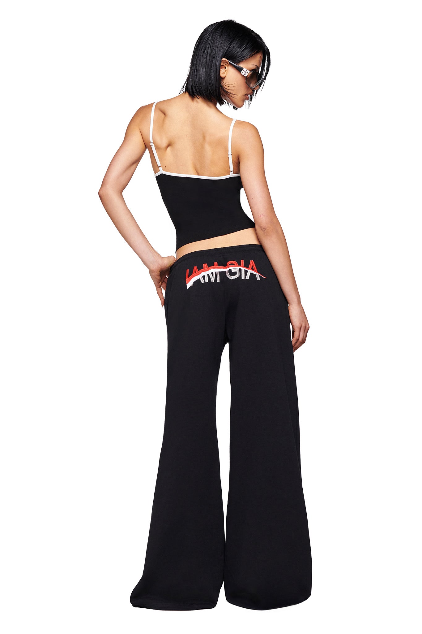 A person in a black strap top and I.AM.GIA's JUSTINE PANT - BLACK, featuring "LAMGIA" in white and red on the back, poses with one hand on their hip, facing away from the camera against a white background.