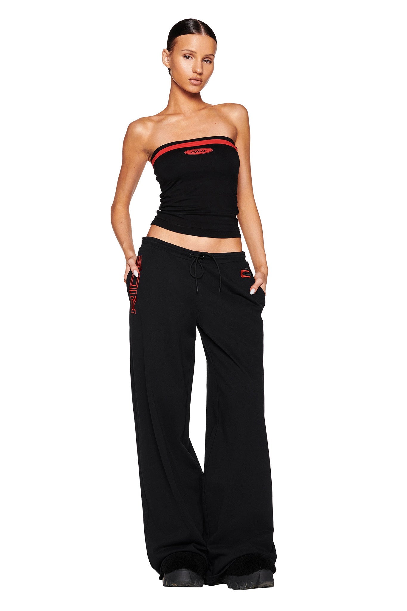 A female model stands against a white background, wearing an I.AM.GIA strapless black top with red trim and lettering, along with the JUSTINE PANT - BLACK from I.AM.GIA, featuring matching red accents. She has one hand in her pocket and a confident expression.