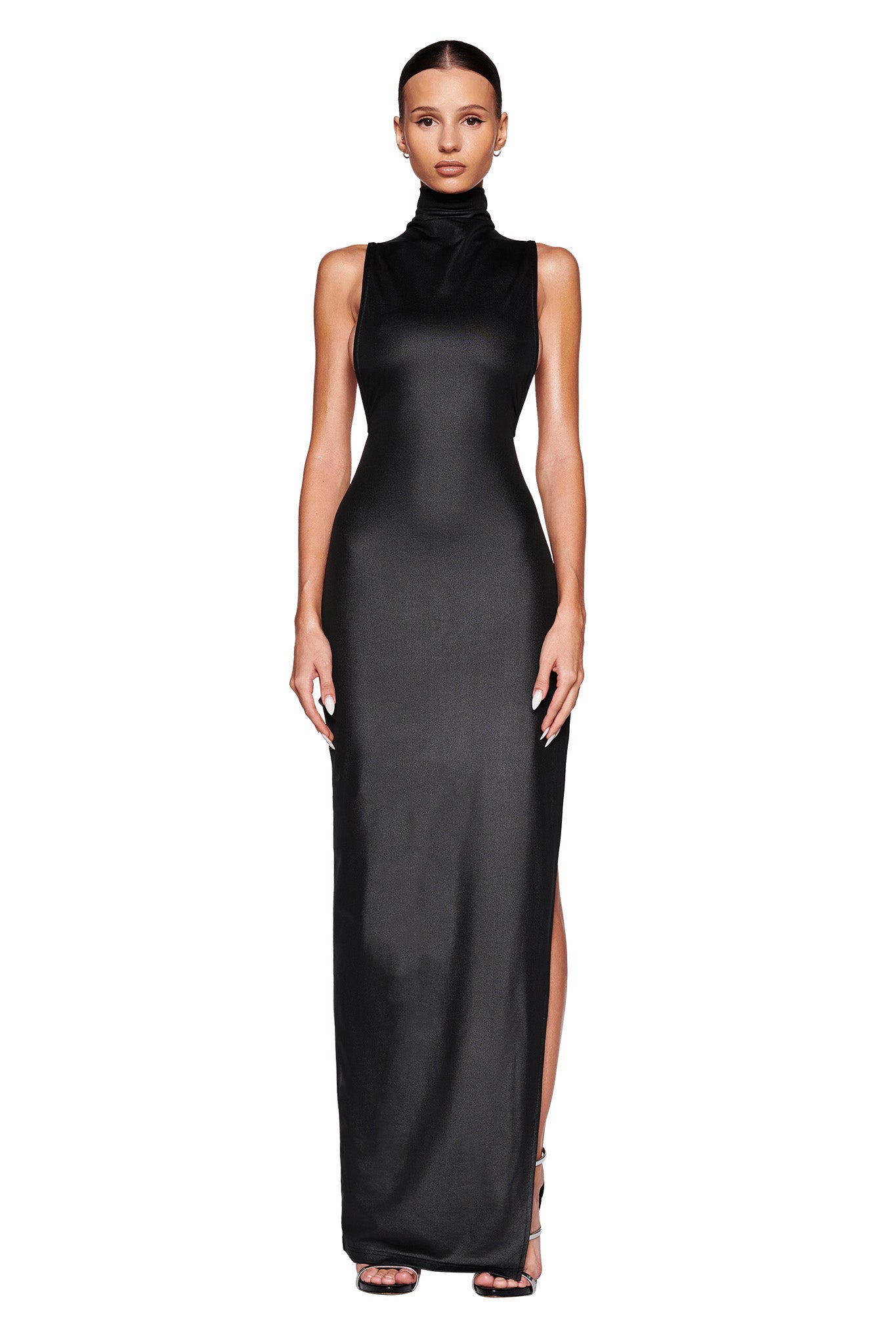 A woman is standing against a white background, wearing the TAGAN MAXI DRESS in black from I.AM.GIA. The gown is sleeveless, high-necked, form-fitting, and reaches the floor. She has her hair pulled back and is wearing black high-heeled shoes. Her arms are relaxed at her sides.