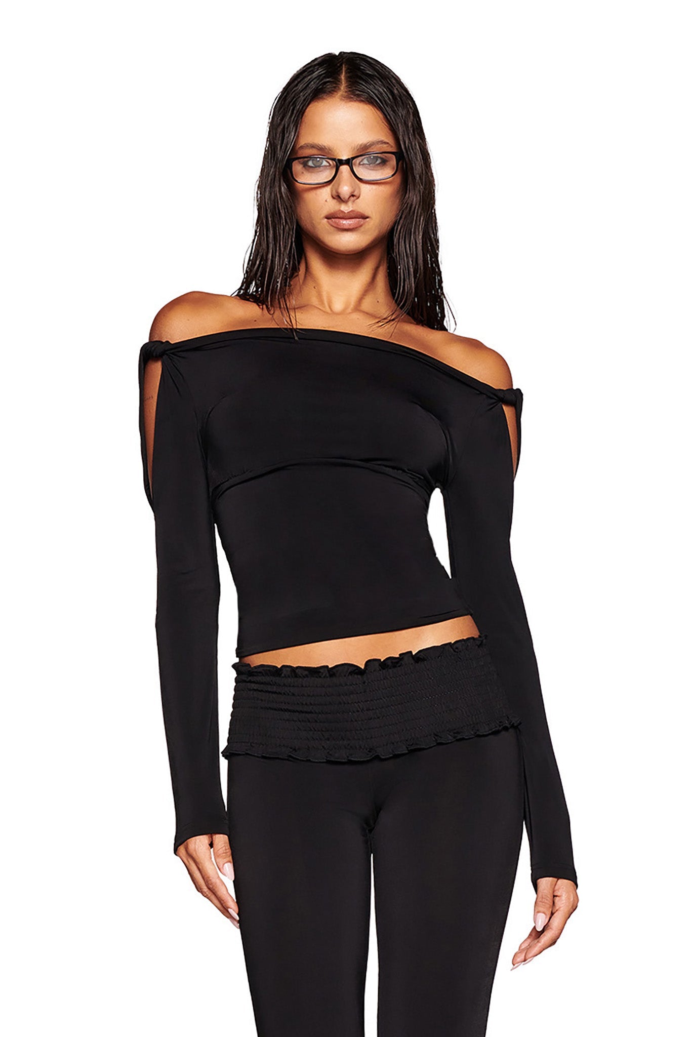 A person with long, wet-looking hair and glasses is wearing a stylish black LORELAI TOP - BLACK by I.AM.GIA and matching black pants. The off-the-shoulder top features cut-out shoulder detailing, while the pants boast a textured waistband. The background is plain white.