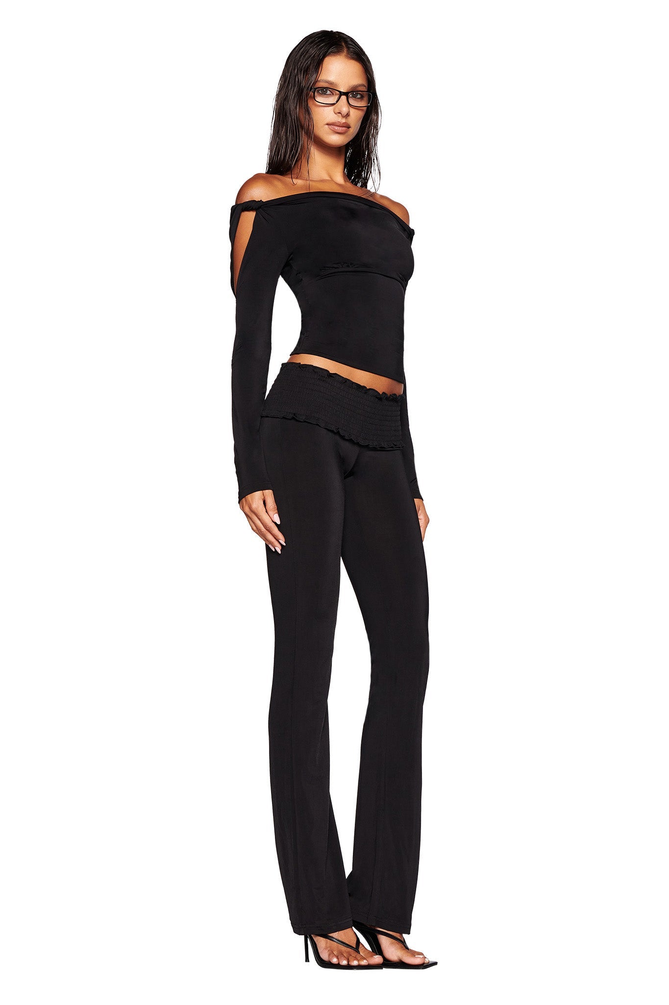 A woman with long, dark hair and glasses stands against a white background, wearing an off-the-shoulder black Lorelai Top by I.AM.GIA and bodycon silhouette pants. She completes her chic look with black high-heeled sandals.