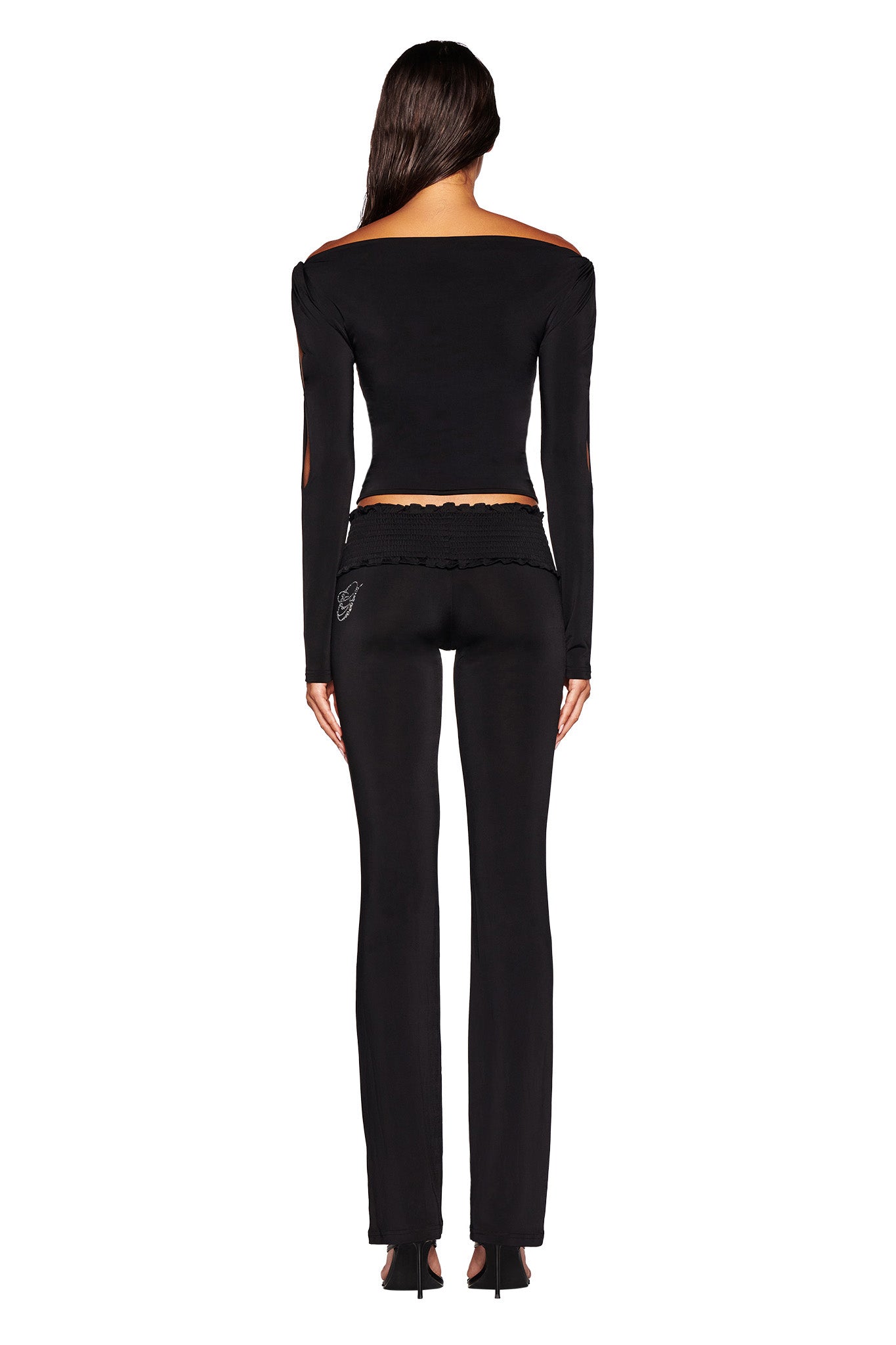 A woman with long, dark hair stands facing away, showcasing the bodycon silhouette of her black LORELAI TOP from I.AM.GIA and matching black pants. The outfit features small cut-out details on the shoulders and upper back, paired with sleek black heels. The background is plain white.