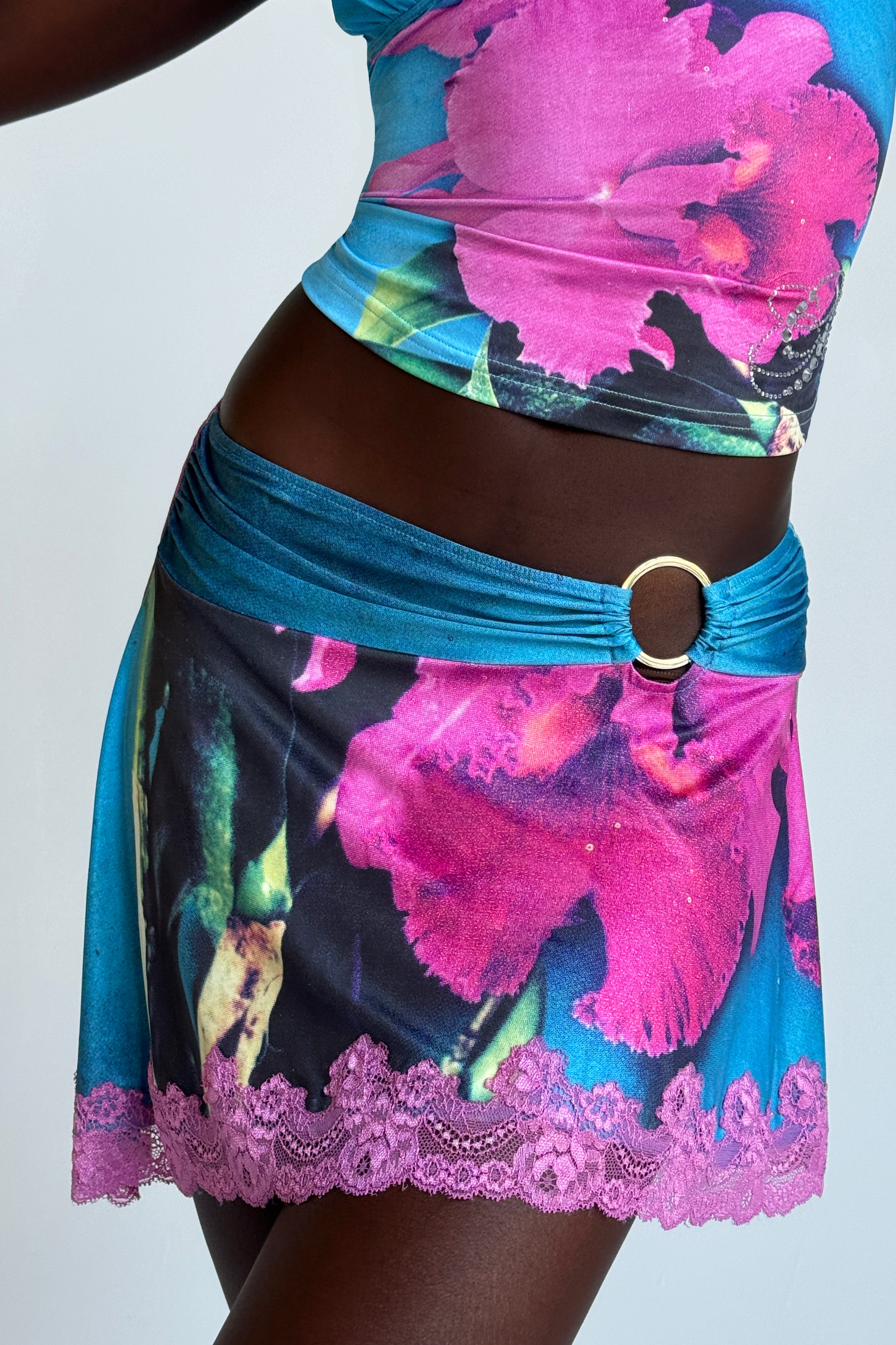 Close-up of a person wearing the ODETTA SKIRT - MULTI from I.AM.GIA, adorned with vibrant pink and blue orchid prints. The outfit features a custom skirt with lace trim at the hem and is complemented by a metal ring accessory at the side. The background is plain light gray.