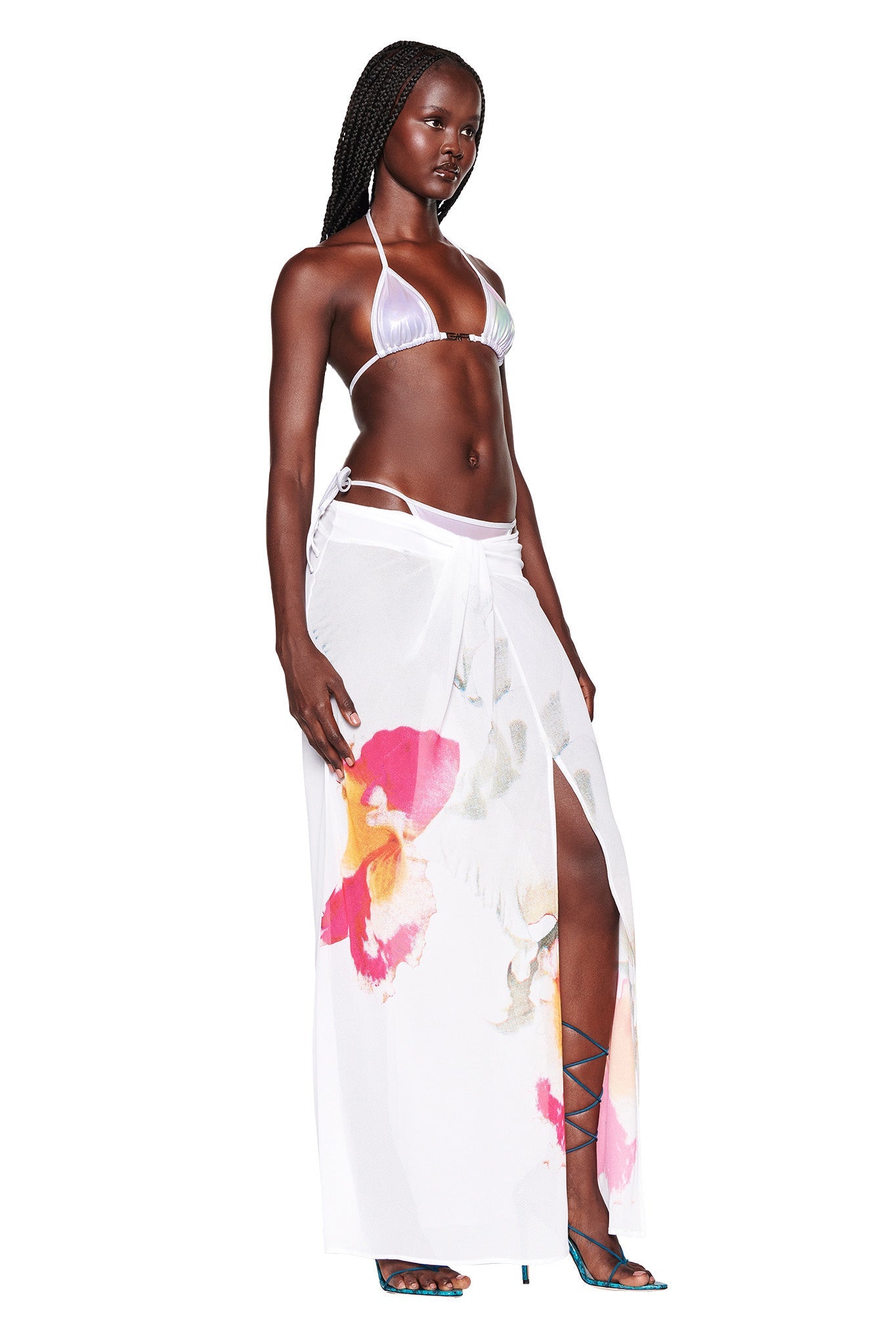 A woman with braided hair is wearing the ALOHI BIKINI TOP in WHITE : PEARL and a sheer white sarong adorned with colorful floral patterns. Complementing her look with strappy blue heels, she stands confidently against a plain white background, embodying the edgy style of I.AM.GIA.