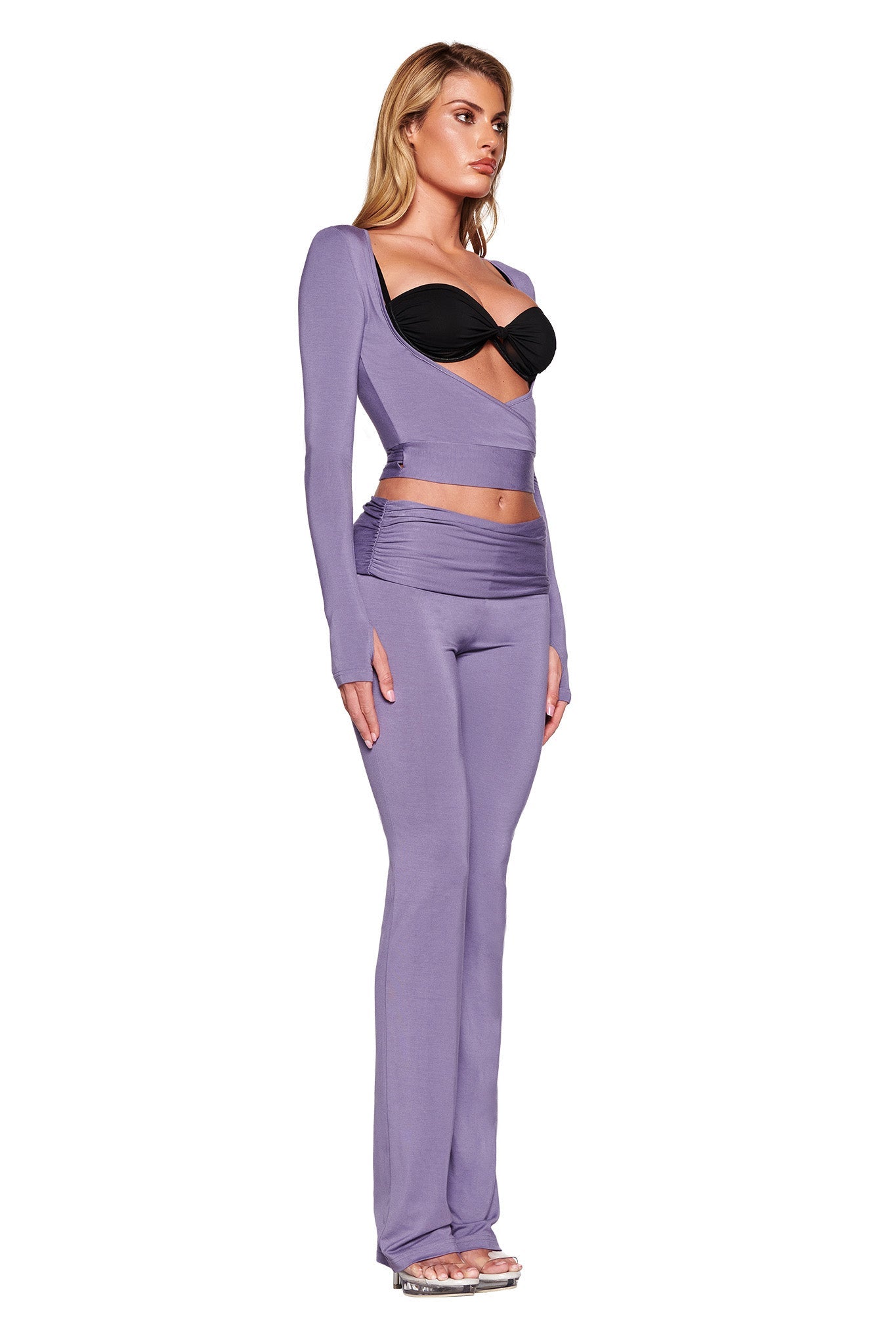 A woman with long blonde hair is standing in a side profile pose. She is wearing the I.AM.GIA STELLA TOP in Dusty Lilac, featuring a black cutout in the front, paired with high-waisted, form-fitting pants made of soft jersey fabrication. The bodycon silhouette highlights her figure, and she completes the look with heels.