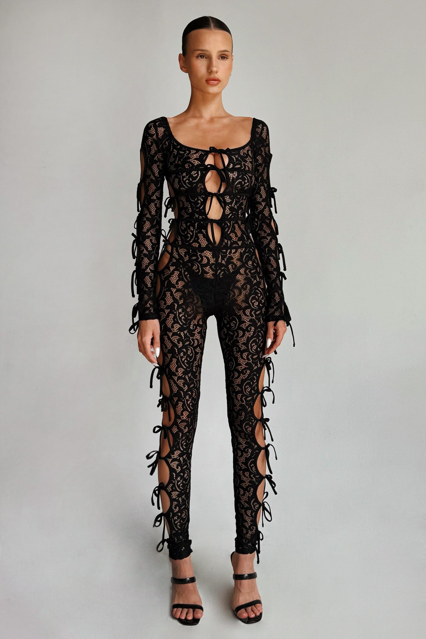 Against a plain backdrop, a woman stands confidently wearing the LYSANDRA CATSUIT - BLACK from I.AM.GIA. The black, lace, long-sleeved bodysuit features multiple cut-outs and tied sections adorned with ribbons. She pairs it with black high-heeled sandals and has her hair slicked back as she gazes at the camera.