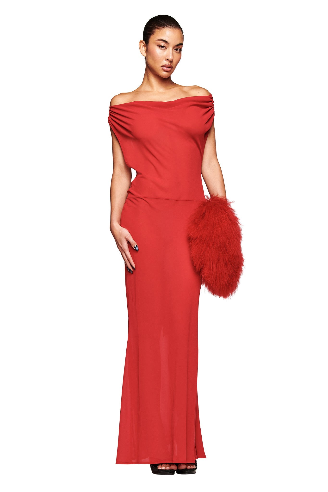 A person poses gracefully against a white background wearing the I.AM.GIA KAHLO MAXI DRESS in red, paired with a large fluffy red clutch. Their dark hair is pulled back, highlighting the dress’s off-the-shoulder design.