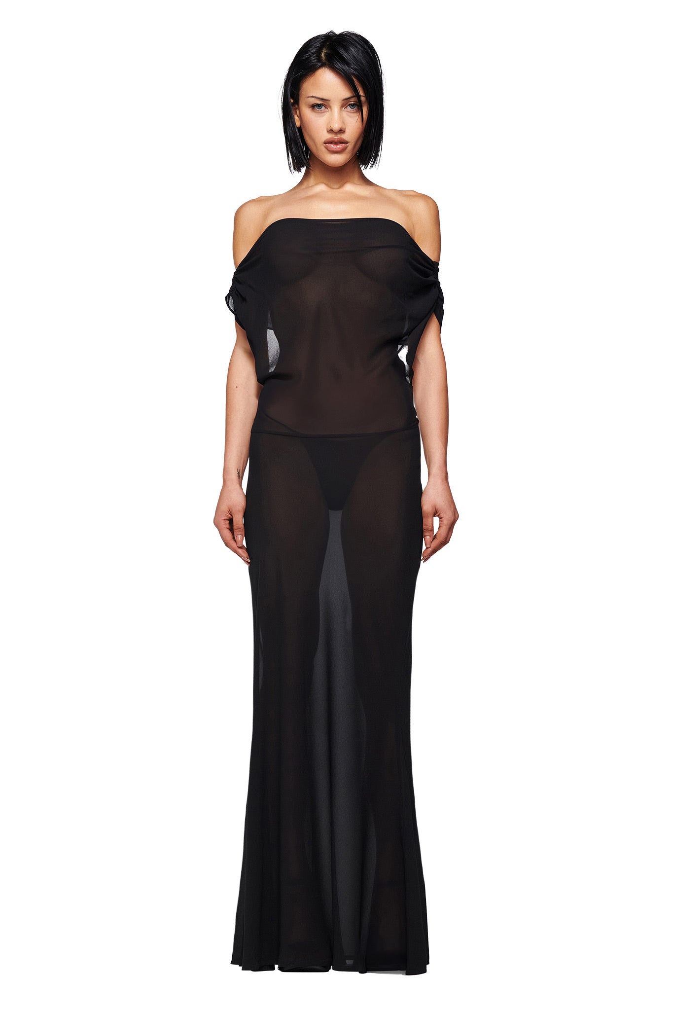 A woman with chin-length dark hair and a neutral expression models the I.AM.GIA KAHLO MAXI DRESS in black—an off-the-shoulder style with a sheer long skirt and visible black undergarments—against a plain white background.