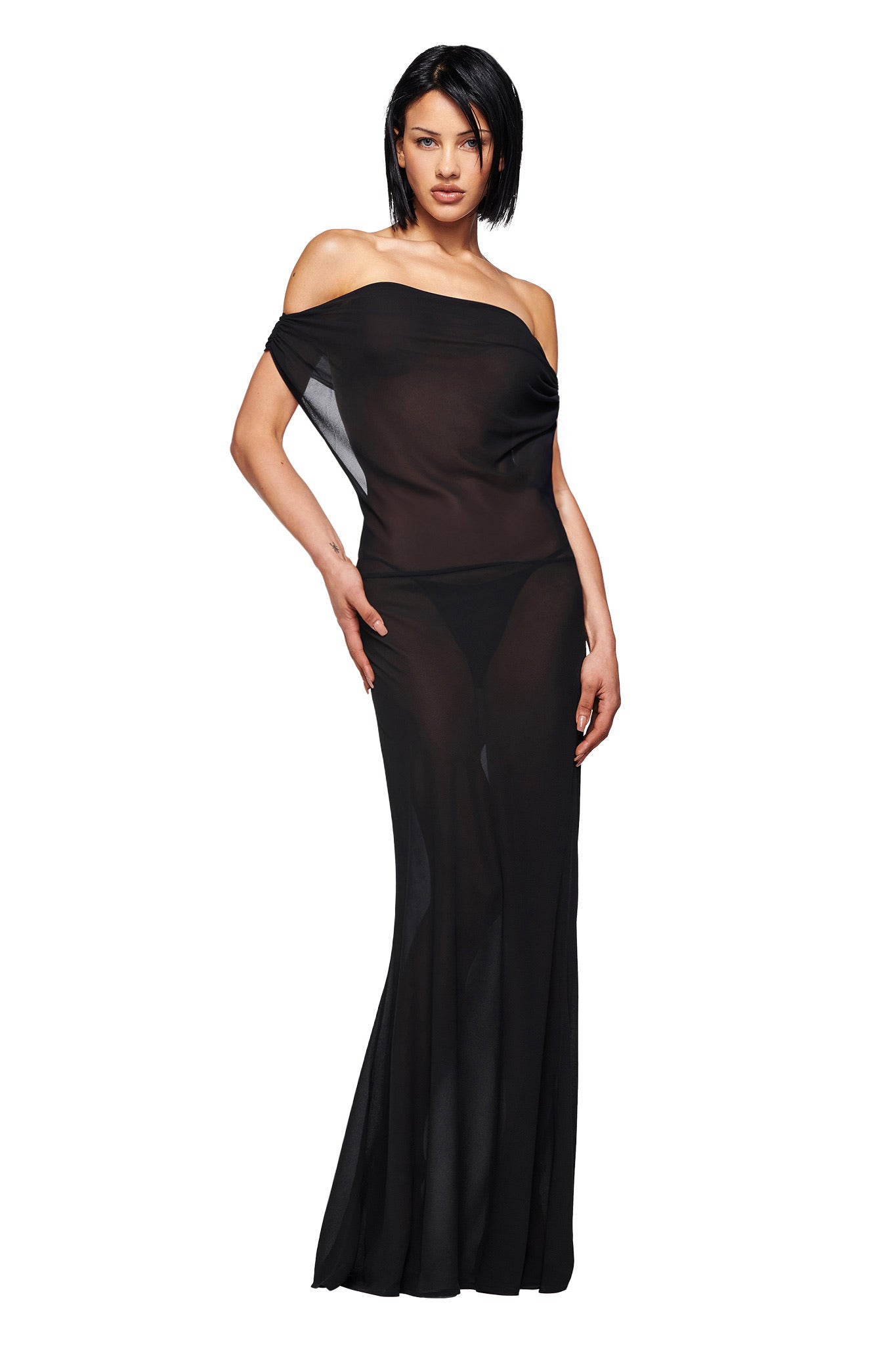 A woman with straight dark hair models the I.AM.GIA KAHLO MAXI DRESS in black—an off-the-shoulder, sheer georgette maxi featuring a long skirt and visible black undergarments. She poses against a white background, one hand on her hip.
