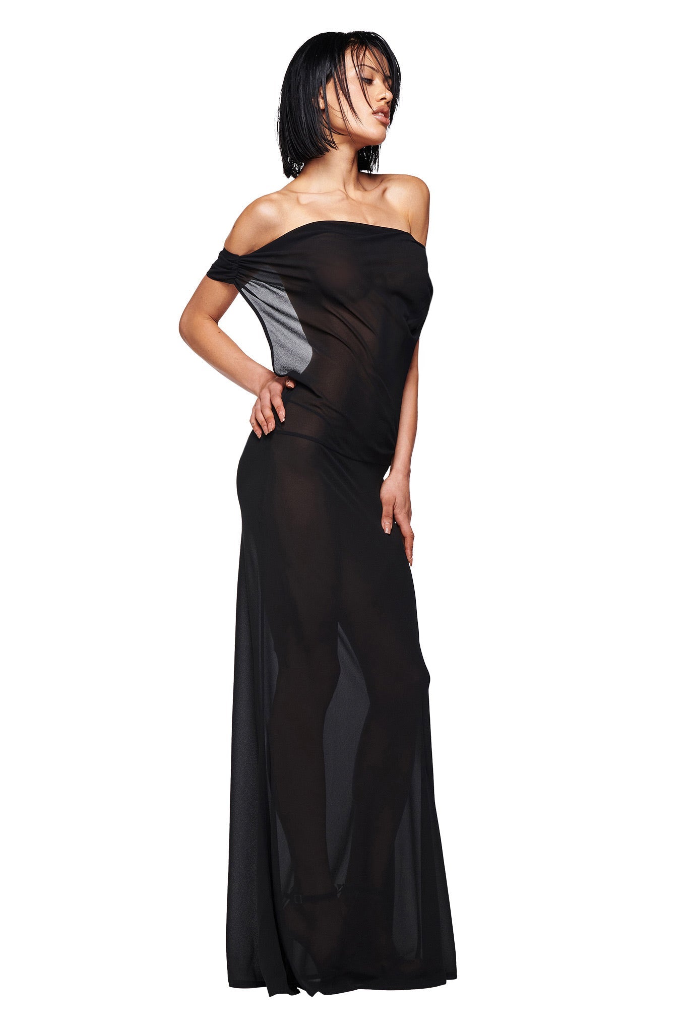 A woman with a short bob wears the I.AM.GIA KAHLO MAXI DRESS in black—a sheer, off-the-shoulder, lightweight georgette gown. She stands with one hand on her hip, gazing to the side against a plain white background.