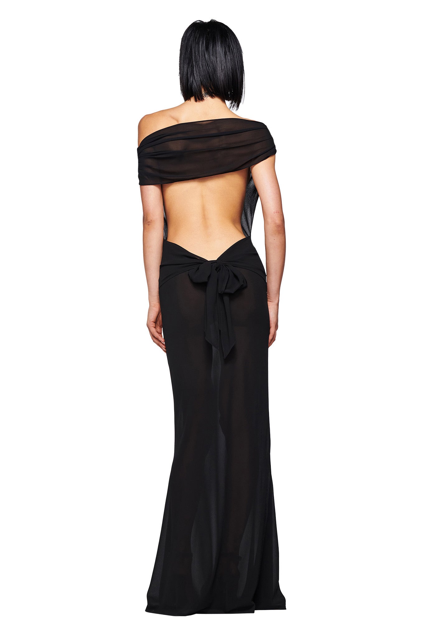 A person with short black hair models the I.AM.GIA KAHLO MAXI DRESS in black, featuring an off-shoulder design, sheer open back, and large waist bow, standing with their back to the camera against a white background.