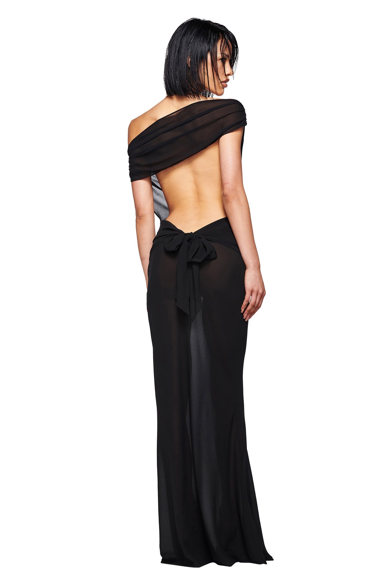 A woman with short black hair models the I.AM.GIA KAHLO MAXI DRESS in black—a lightweight, off-the-shoulder georgette gown with a backless, floor-length design and a large bow at the lower back—against a white background.