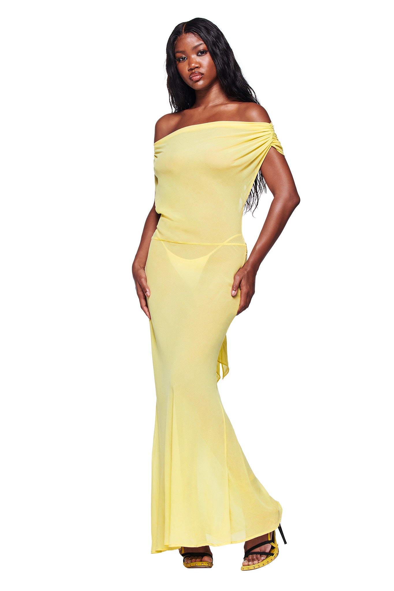 A woman models the I.AM.GIA KAHLO MAXI DRESS in yellow, striking a pose with one hand on her hip. She pairs the off-the-shoulder, flowing dress with black heeled sandals against a white background.