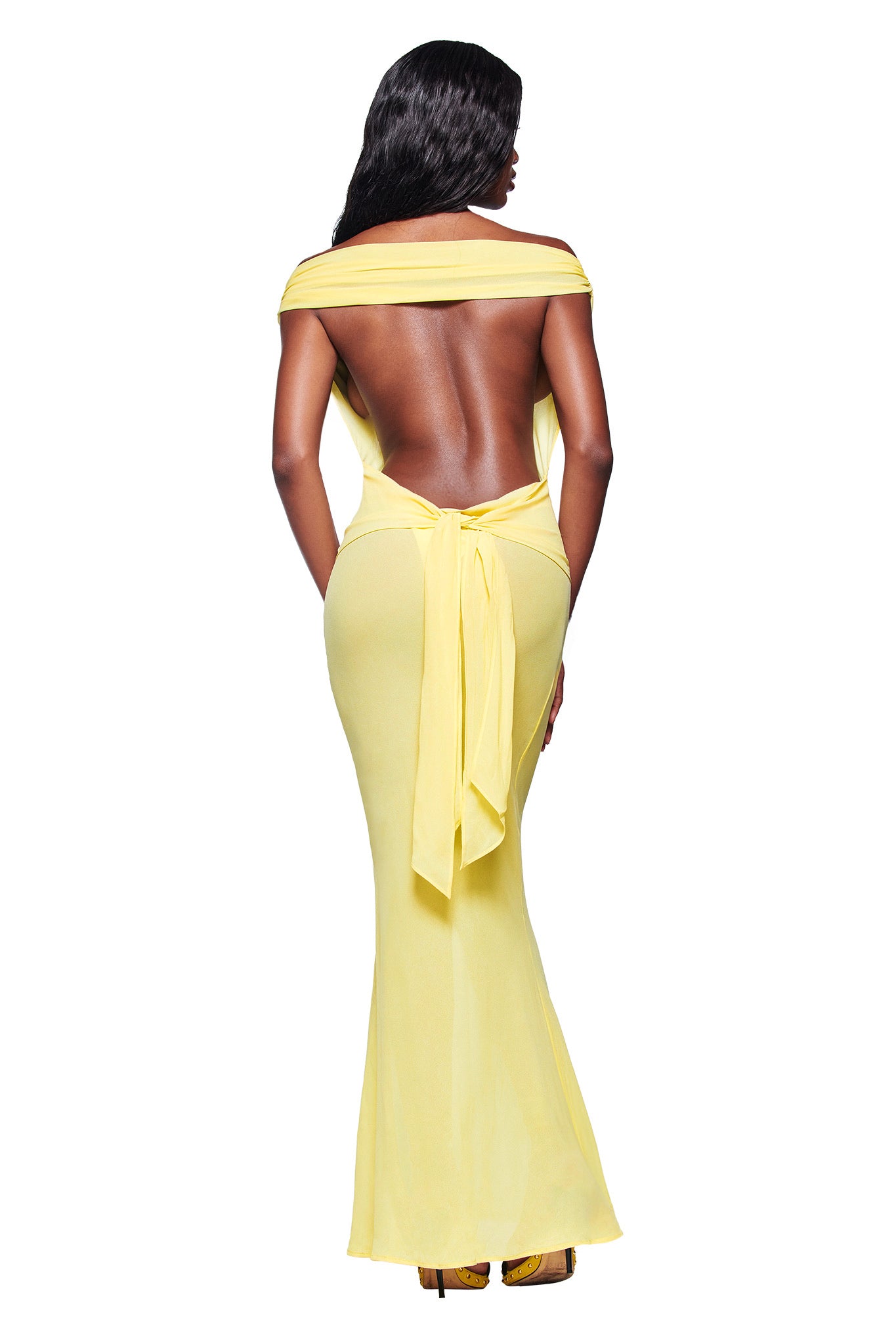 A woman with loose waves and high heels faces away, wearing the I.AM.GIA KAHLO MAXI DRESS in yellow—an off-the-shoulder, backless design with a sash at the waist. The background is white.