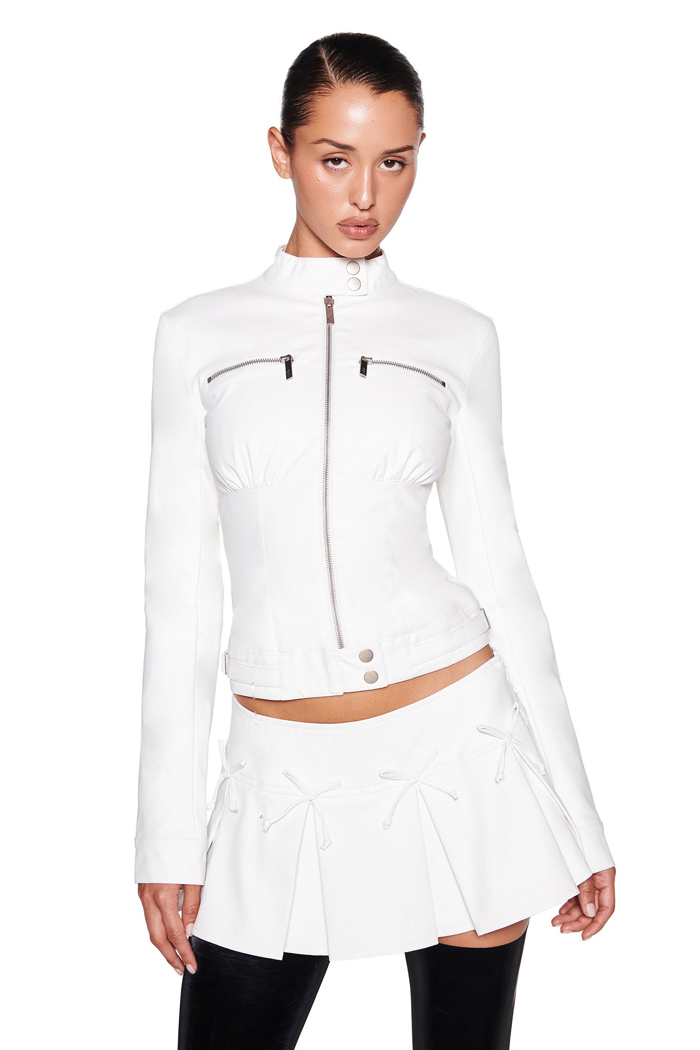 A person dressed in the I.AM.GIA EVERETTE JACKET - WHITE, featuring a sleek, tailored fit and textured faux leather with chest pockets, pairs it with a white pleated skirt. They stand against a white background with dark hair pulled back and pose with their hands slightly forward.