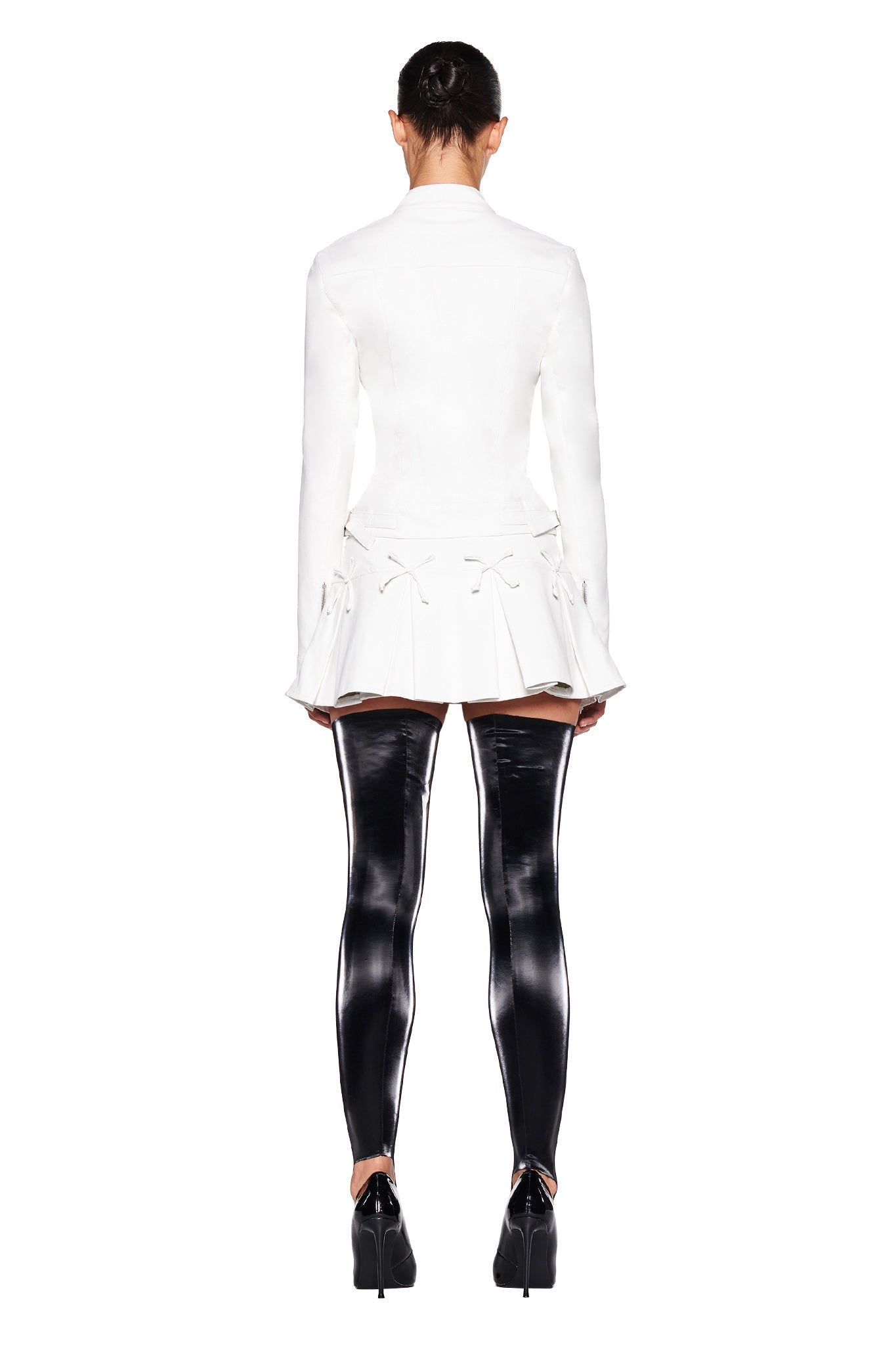 A person is standing with their back to the camera. They are wearing the EVERETTE JACKET in white by I.AM.GIA, a textured faux leather jacket with a tailored fit, complemented by a pleated skirt and black, shiny, thigh-high boots with high heels. Their dark hair is tied up against a plain white background.
