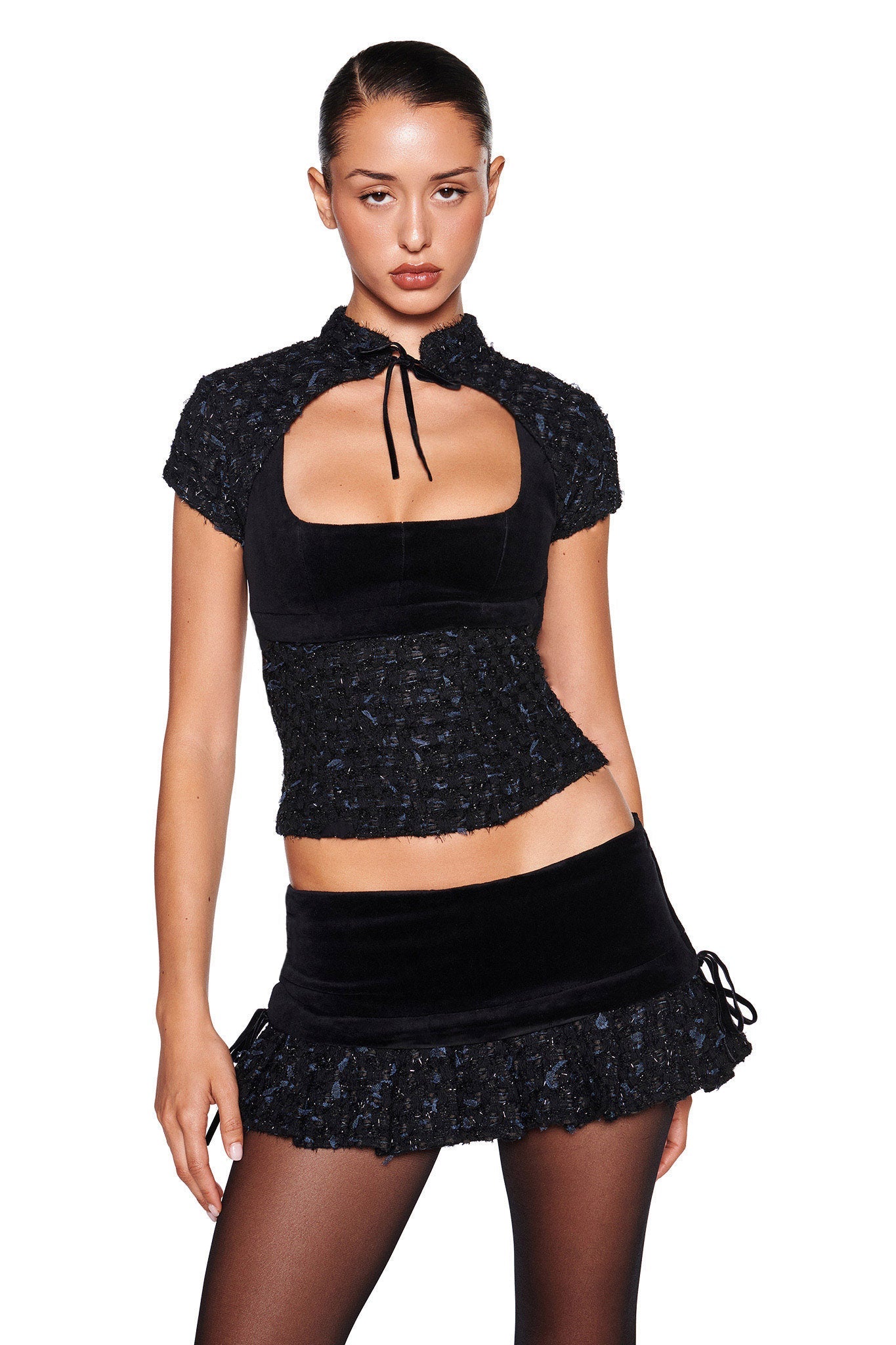 A woman with slicked-back hair stands confidently in the I.AM.GIA SAFFRON 3 PIECE CORSET - BLACK, paired with a matching boucle skirt featuring ruffle details. She completes her look with sheer black tights, posing against a plain white background.