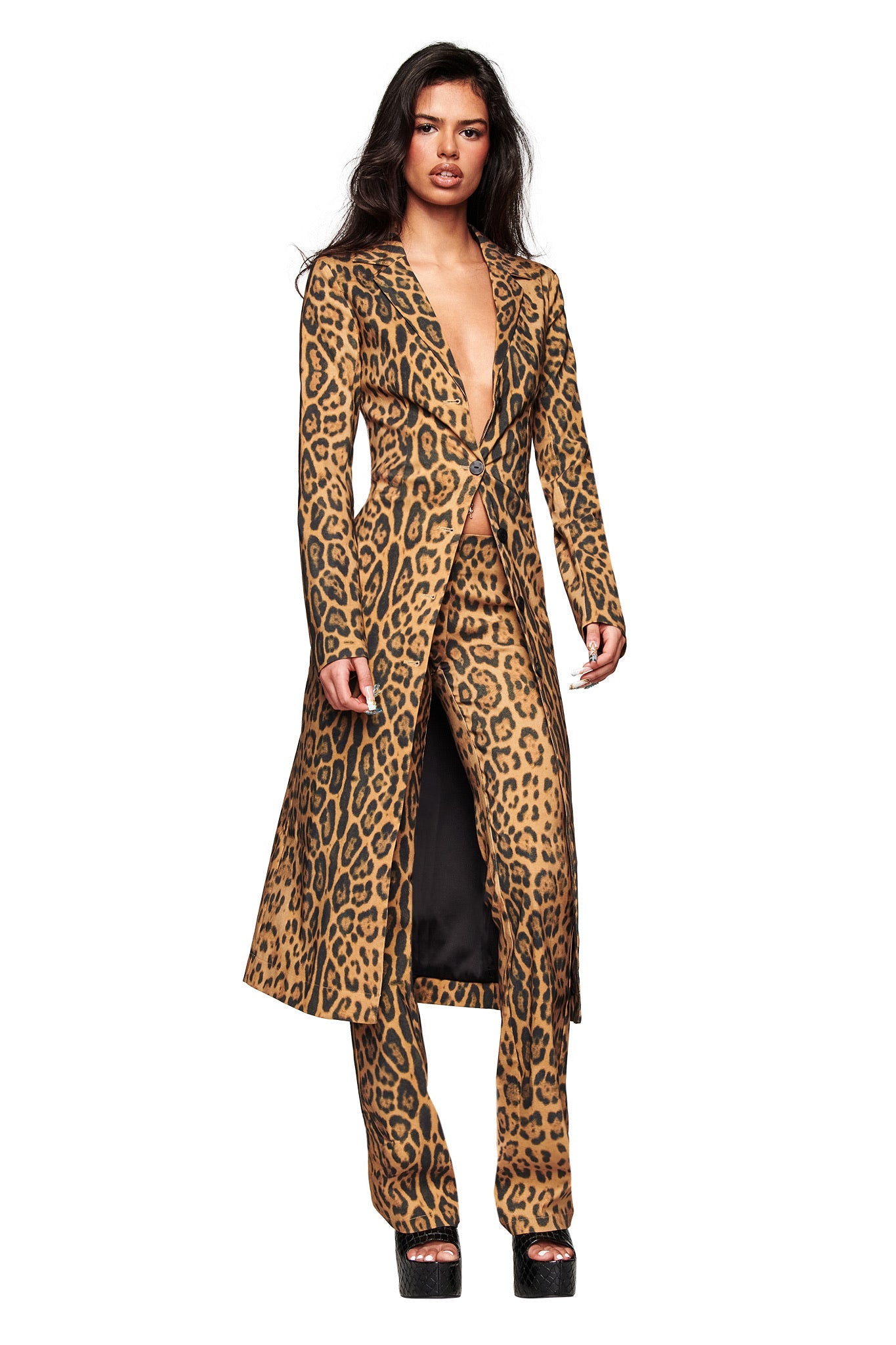 A woman stands confidently in the I.AM.GIA UMA COAT - PANTHERA, styled with matching pants and black platform shoes. The longline animal print coat is open at the neckline as she poses against a white background.