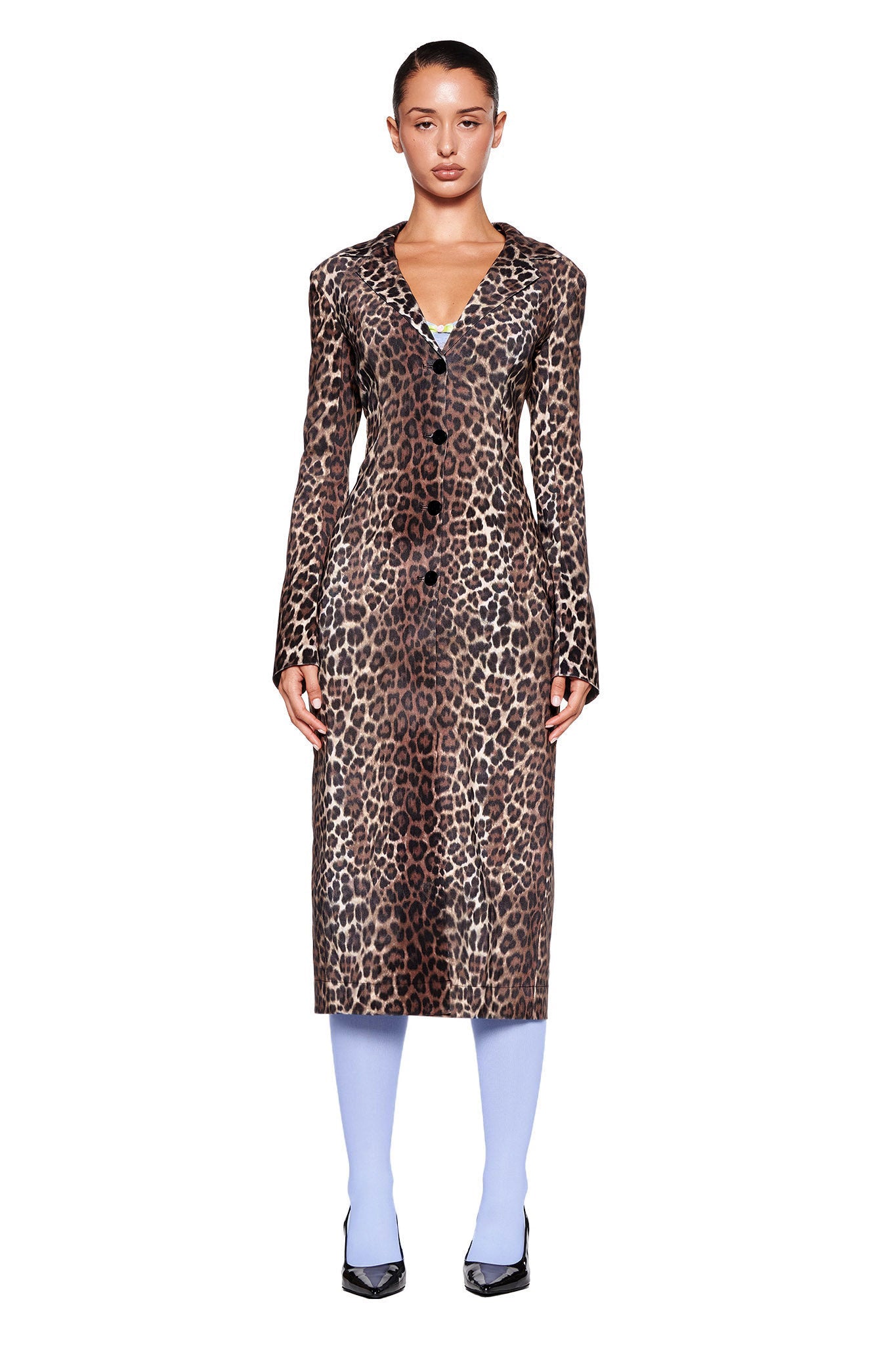 A person stands against a white background wearing the UMA COAT - NEUTRAL : LEOPARD from I.AM.GIA, paired with blue tights and black pointed-toe heels. Their hair is tied back, and they look directly at the camera with a neutral expression.