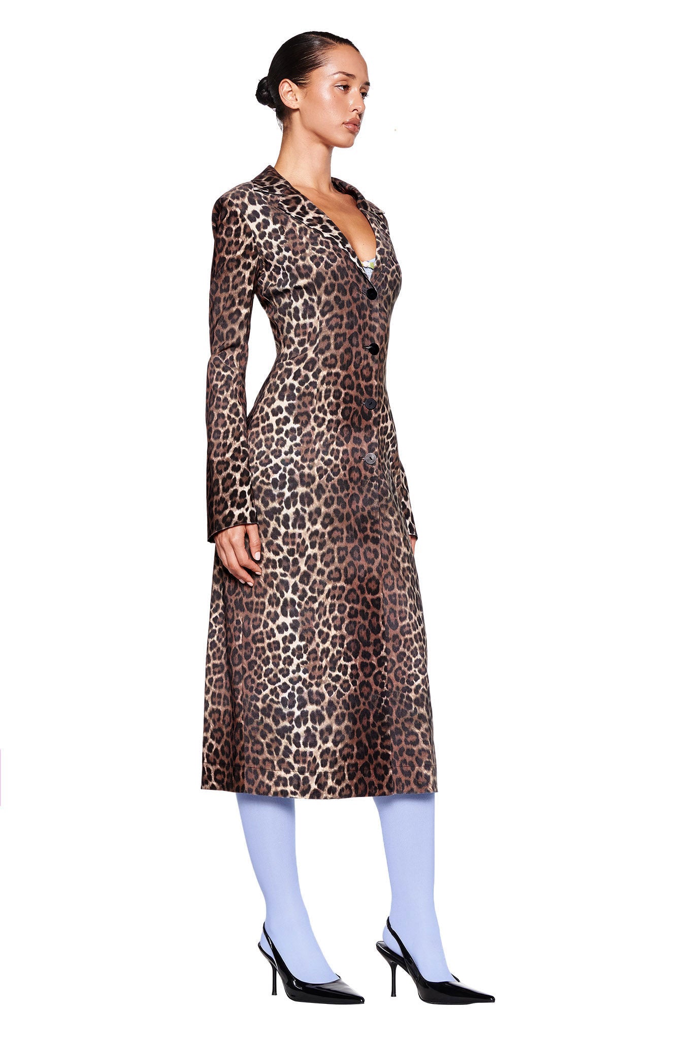 A woman is posing in a side view, wearing the UMA COAT - NEUTRAL : LEOPARD from I.AM.GIA, paired with light blue tights and black pointed-toe heels. Her hair is styled in a sleek bun as she stands against a plain white background, giving off major I.AM.GIA vibes.