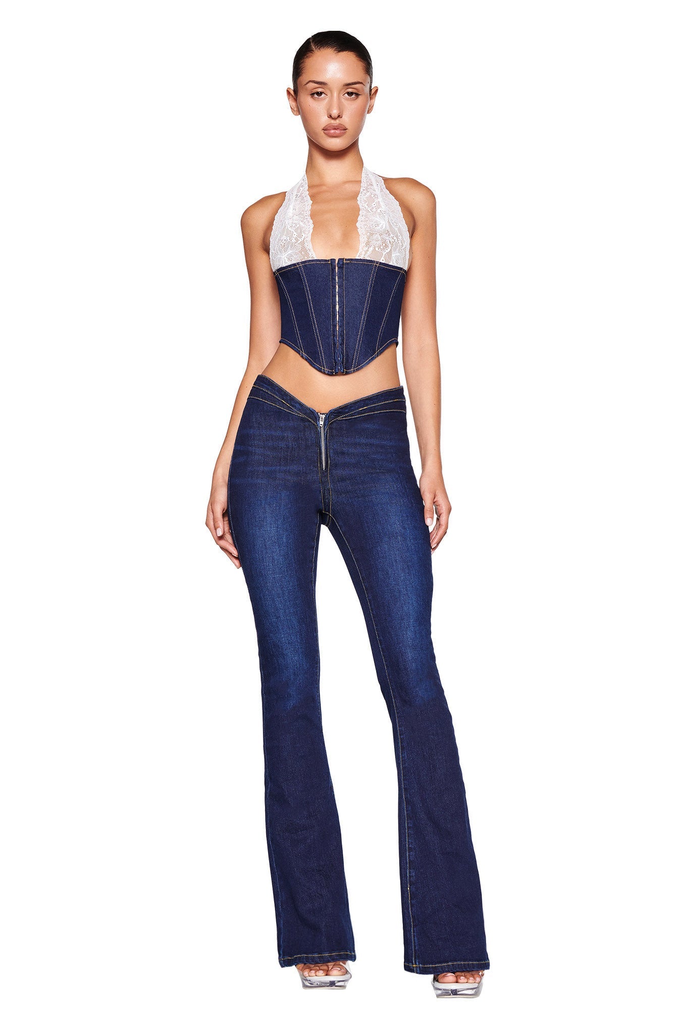 A woman stands against a white background wearing the ELLENA JEAN 2.0 - BLUE: DARK WASH from I.AM.GIA, featuring a dark denim halter top with white lace accents and wide-leg pants that create a flared silhouette. She pairs the outfit with clear high-heeled sandals.