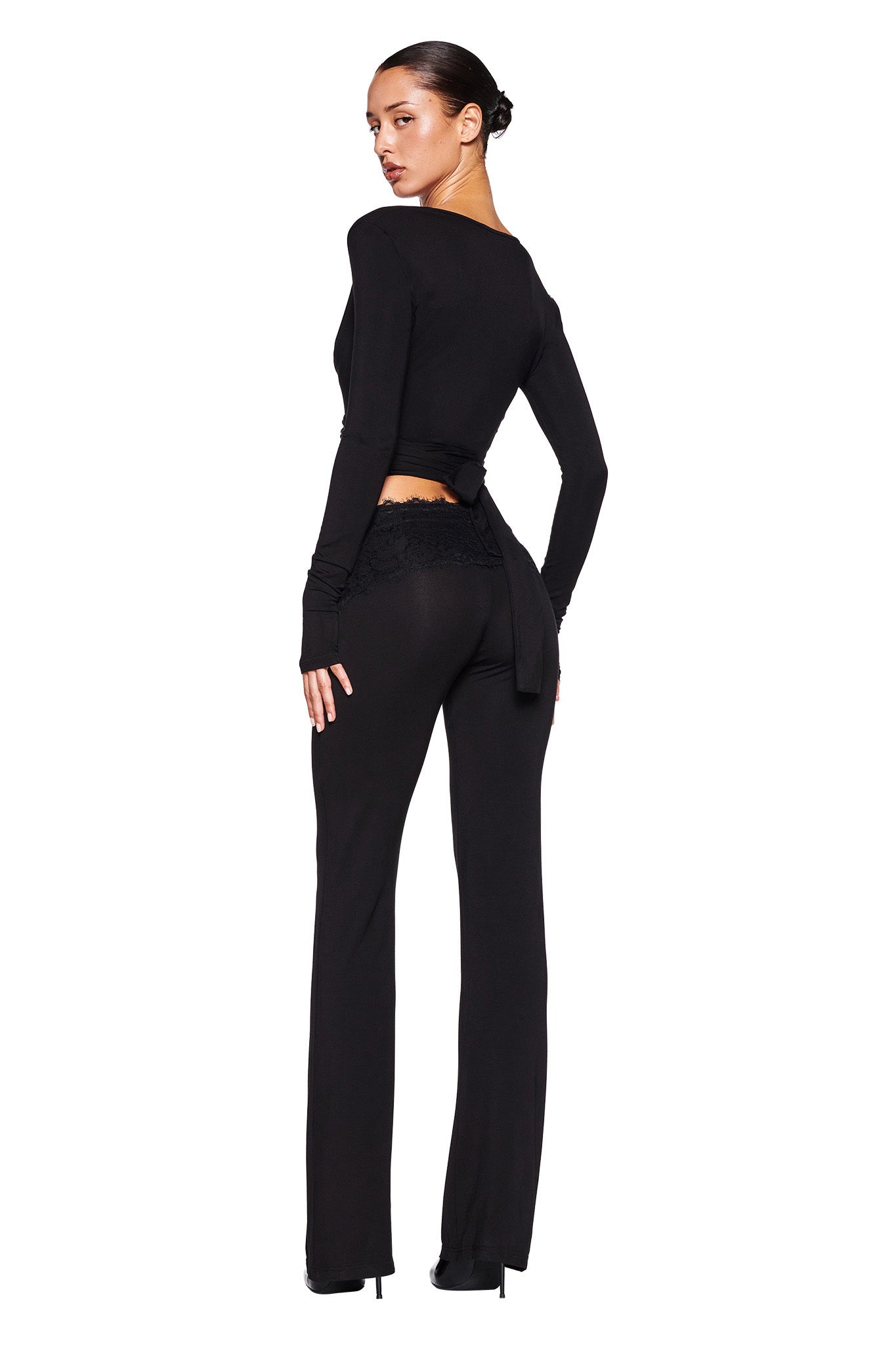 A woman stands facing backward, looking over her left shoulder. She is wearing the MANDY TOP - BLACK by I.AM.GIA, a fitted long-sleeved black wrap top with an open back tied at the waist, paired with matching black pants featuring lace detailing at the waistband. Her hair is styled in a sleek bun, and she's wearing black high heels.