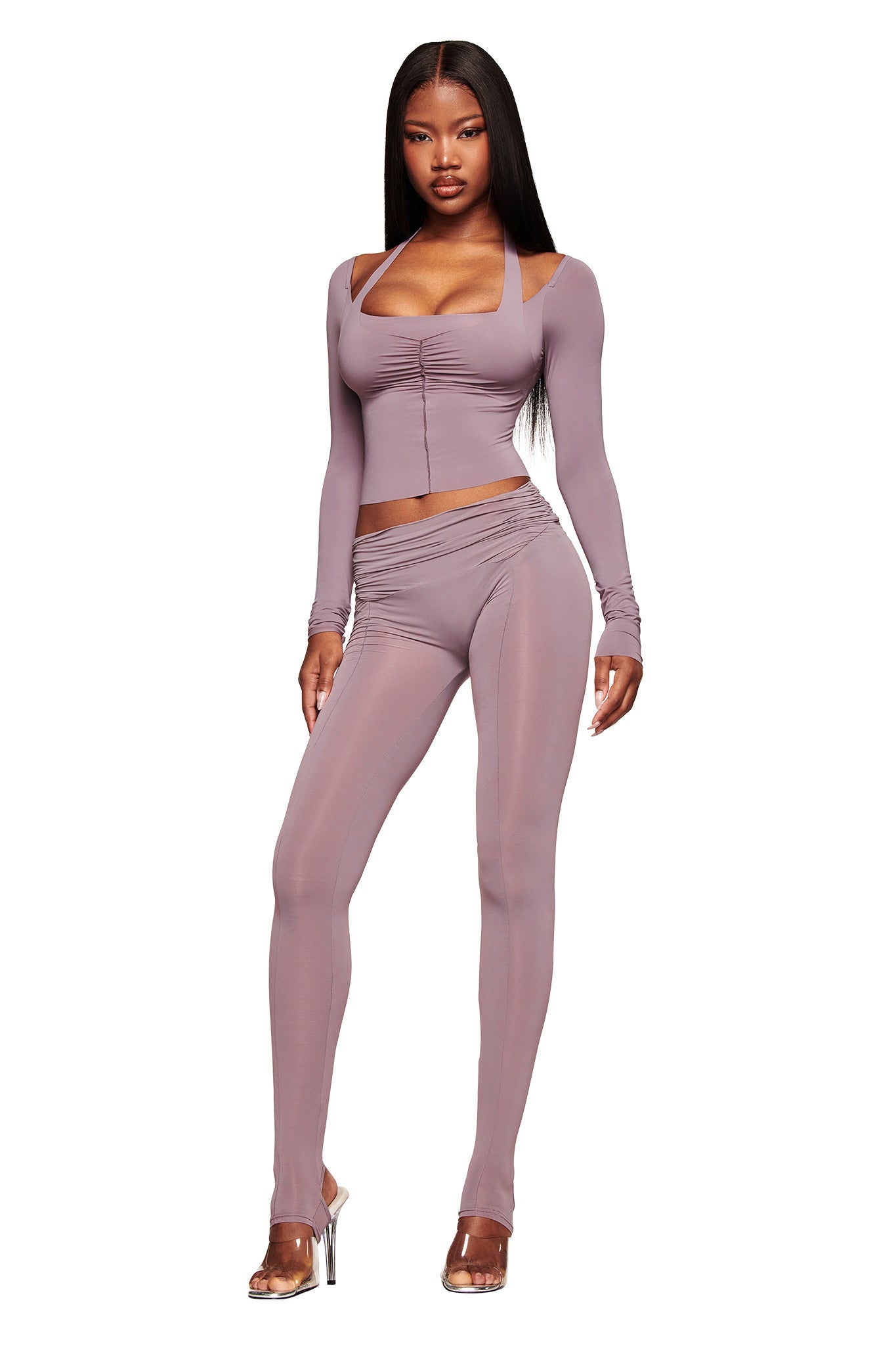 A woman stands confidently in the I.AM.GIA NERINA LEGGING in Dusty Lilac, styled with a mauve long-sleeve crop top. She has long straight hair and wears clear high-heeled shoes. The leggings feature sheer panels and ruched details.