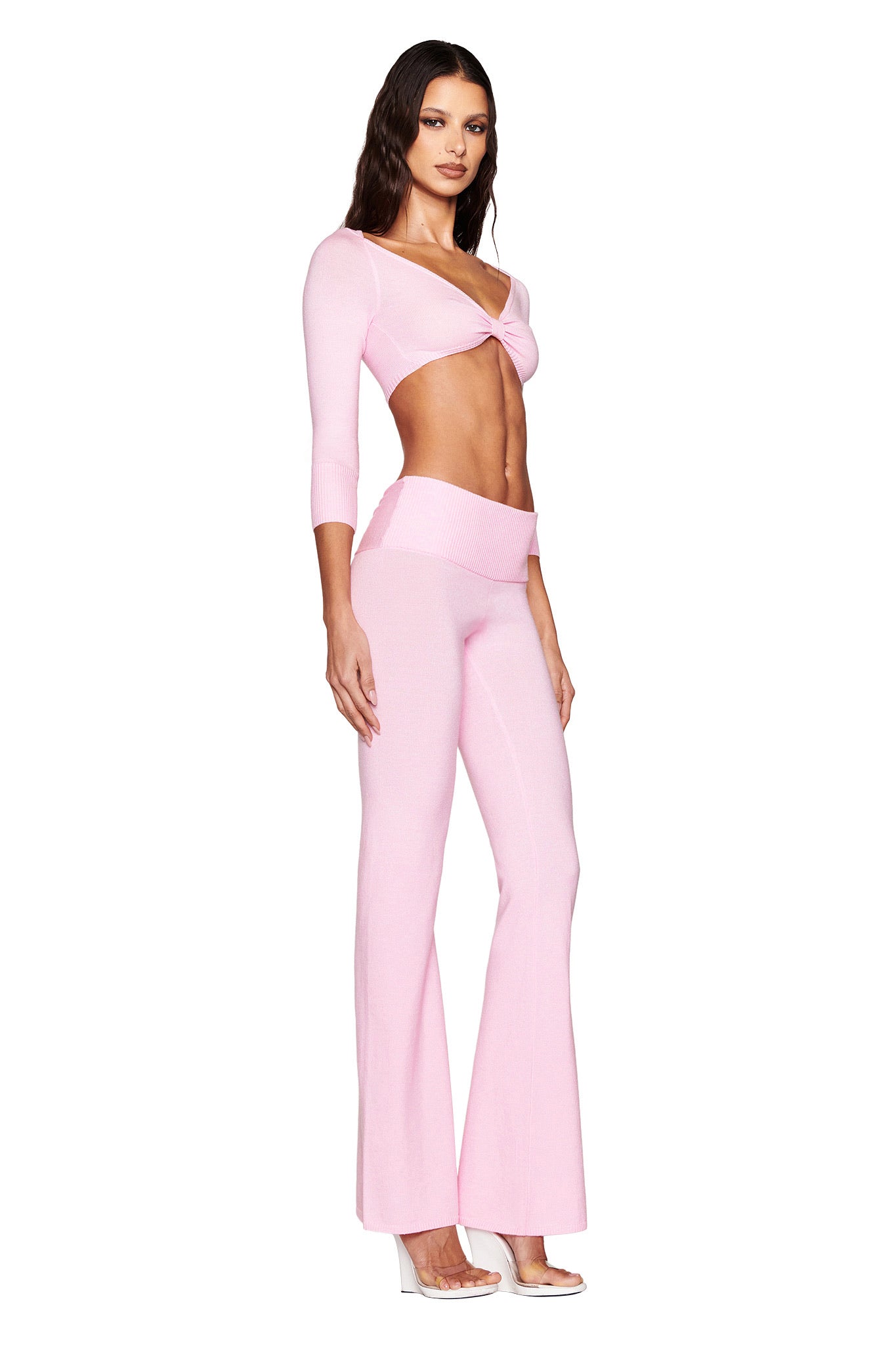 Against a white backdrop, a woman wears I.AM.GIA's MORGANA CROP TOP in pink—a light pink, ultra-soft knit two-piece outfit with flared pants. The top features long sleeves and a twisted front. She lets her hair down and pairs the look with clear high heels.