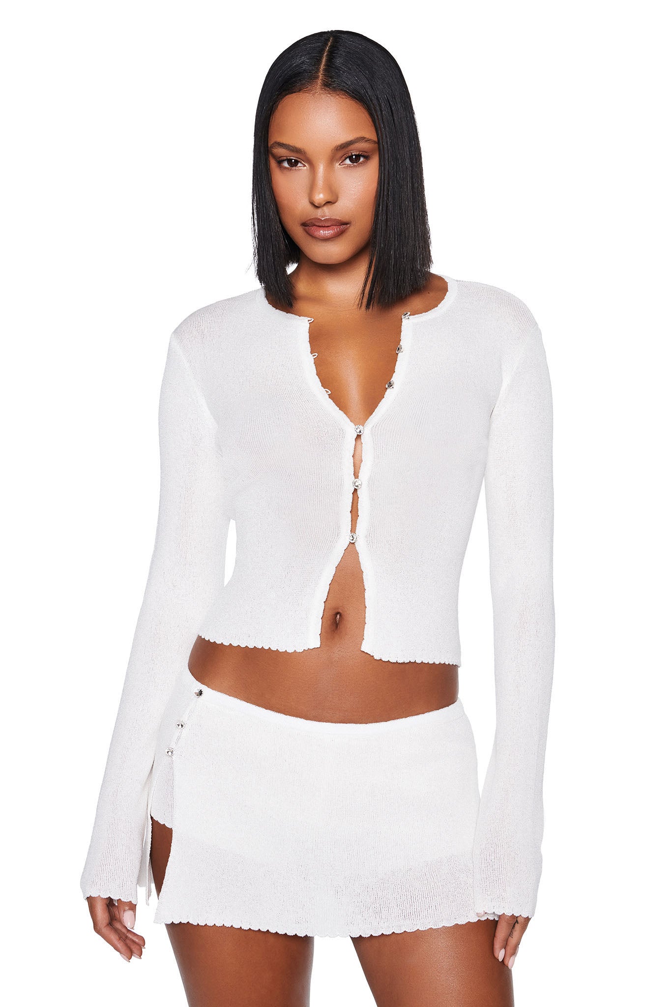 A person wearing the I.AM.GIA SNOW WHITE BLISS SKORT, featuring a white long-sleeve cardigan with scalloped edges and a front closure, paired with the matching mini skort. The outfit is crafted from shimmery lurex yarn, giving it a textured appearance with a subtle sheen. The plain white background enhances its elegance.