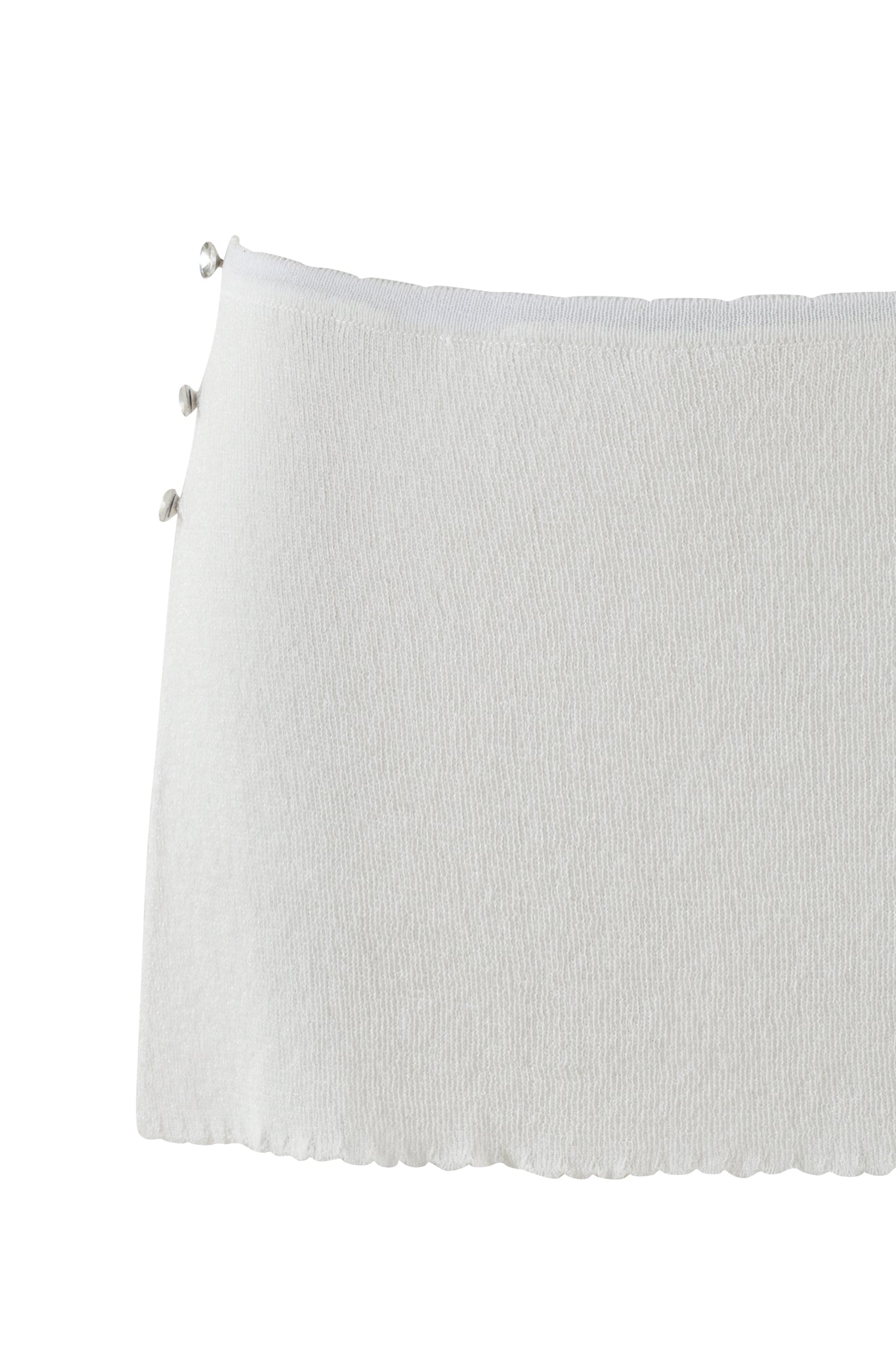 A close-up image of the BLISS SKORT in Snow White from I.AM.GIA shows a horizontally oriented white knitted fabric made from shimmery lurex yarn. The top edge features a scalloped trim with three small loops and button details on the side, adding elegance to this soft, lightweight design.