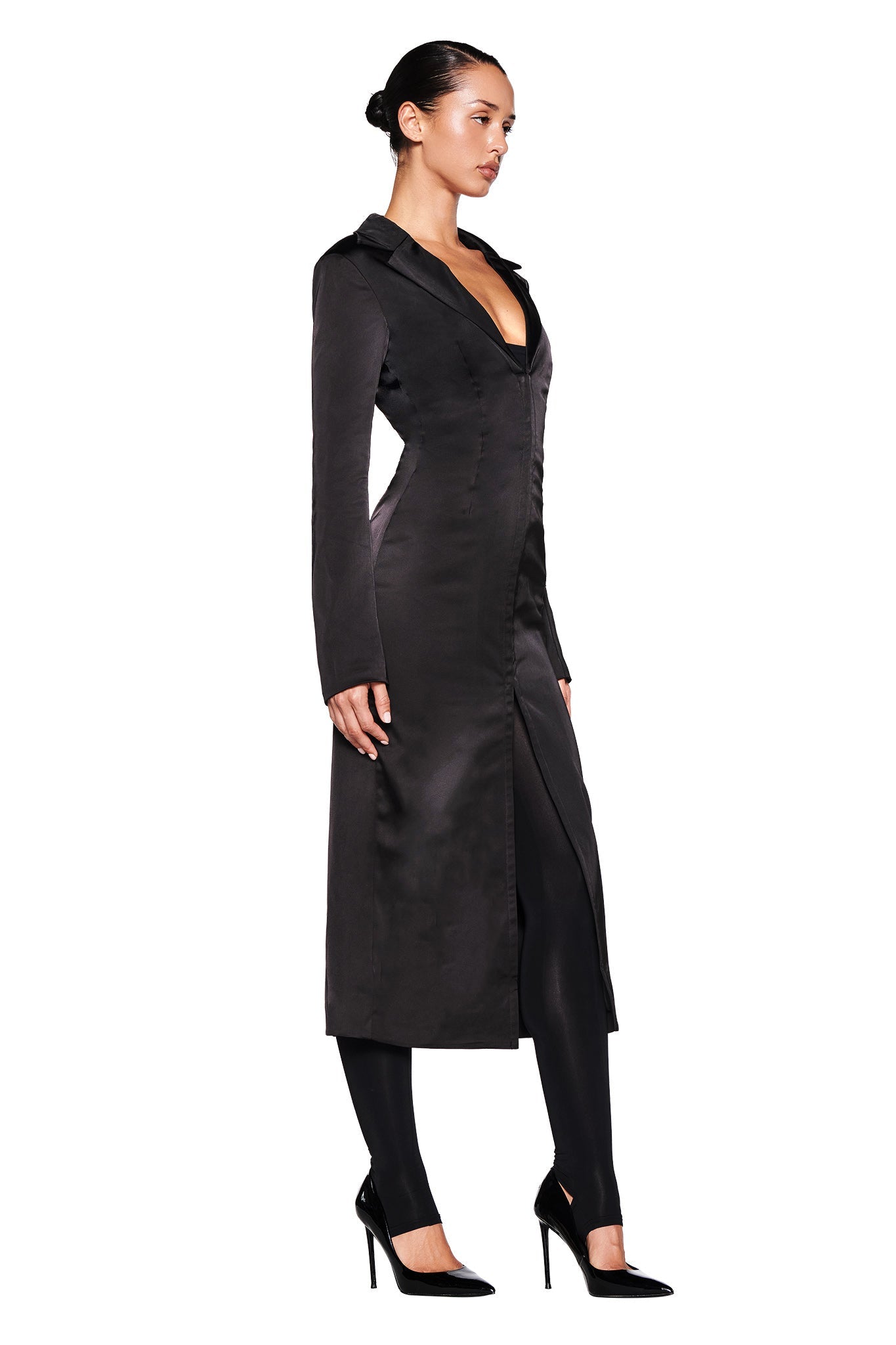 A woman stands in profile against a white background, wearing the I.AM.GIA RAENA COAT in black. The long jacket, featuring a high collar and deep V-neck, is meticulously tailored for a perfect fit. Paired with black leggings and pointed-toe high heels, her sleek bun adds to the sophisticated look of the satin fabrication.