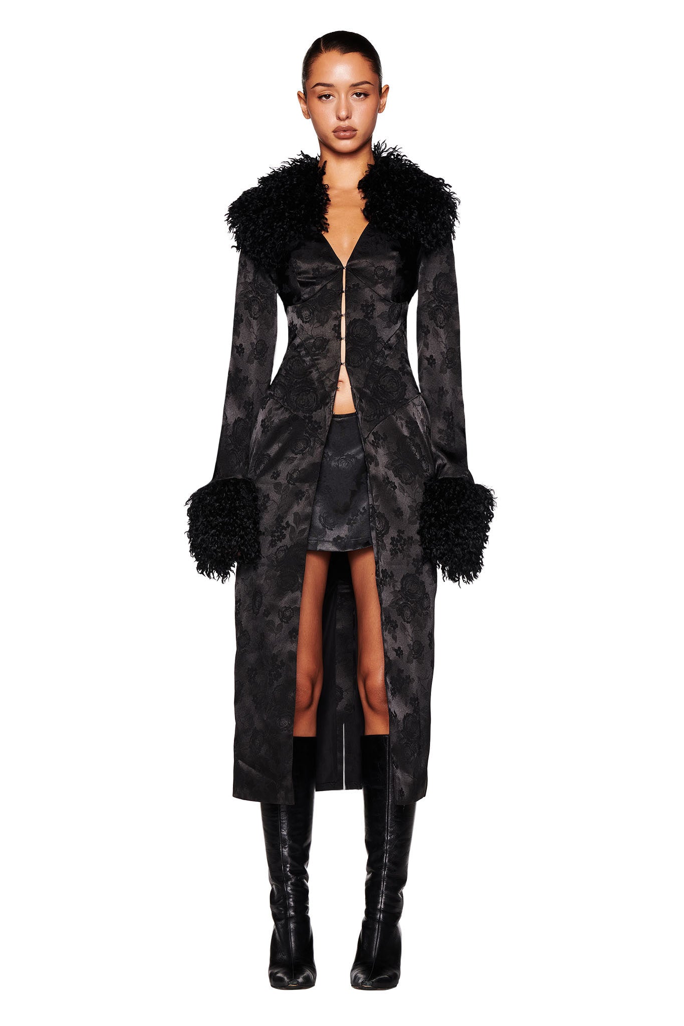 A person is showcasing the I.AM.GIA TAMERA COAT in black, a form-fitting piece featuring a floral jacquard design and accented with faux Mongolian fur on the collar and cuffs. They complement it with a short skirt and knee-high black boots while posing against a white background.
