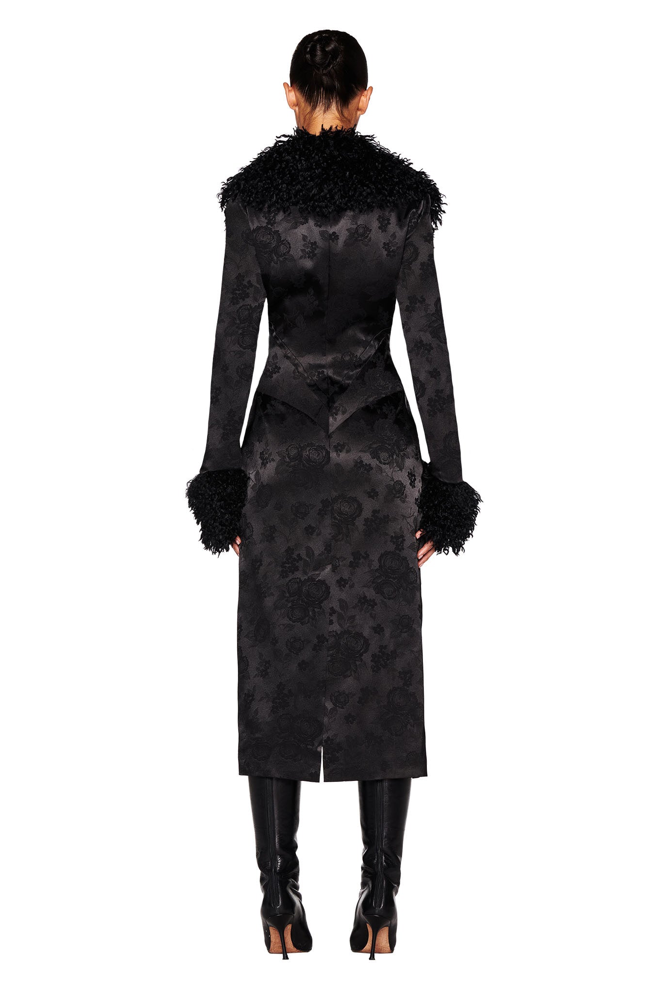 A person is standing with their back to the camera, elegantly draped in the black TAMERA COAT by I.AM.GIA, which features a floral jacquard design and textured sleeves. Their ensemble is complete with black knee-high boots against a white backdrop, creating an exquisite silhouette.
