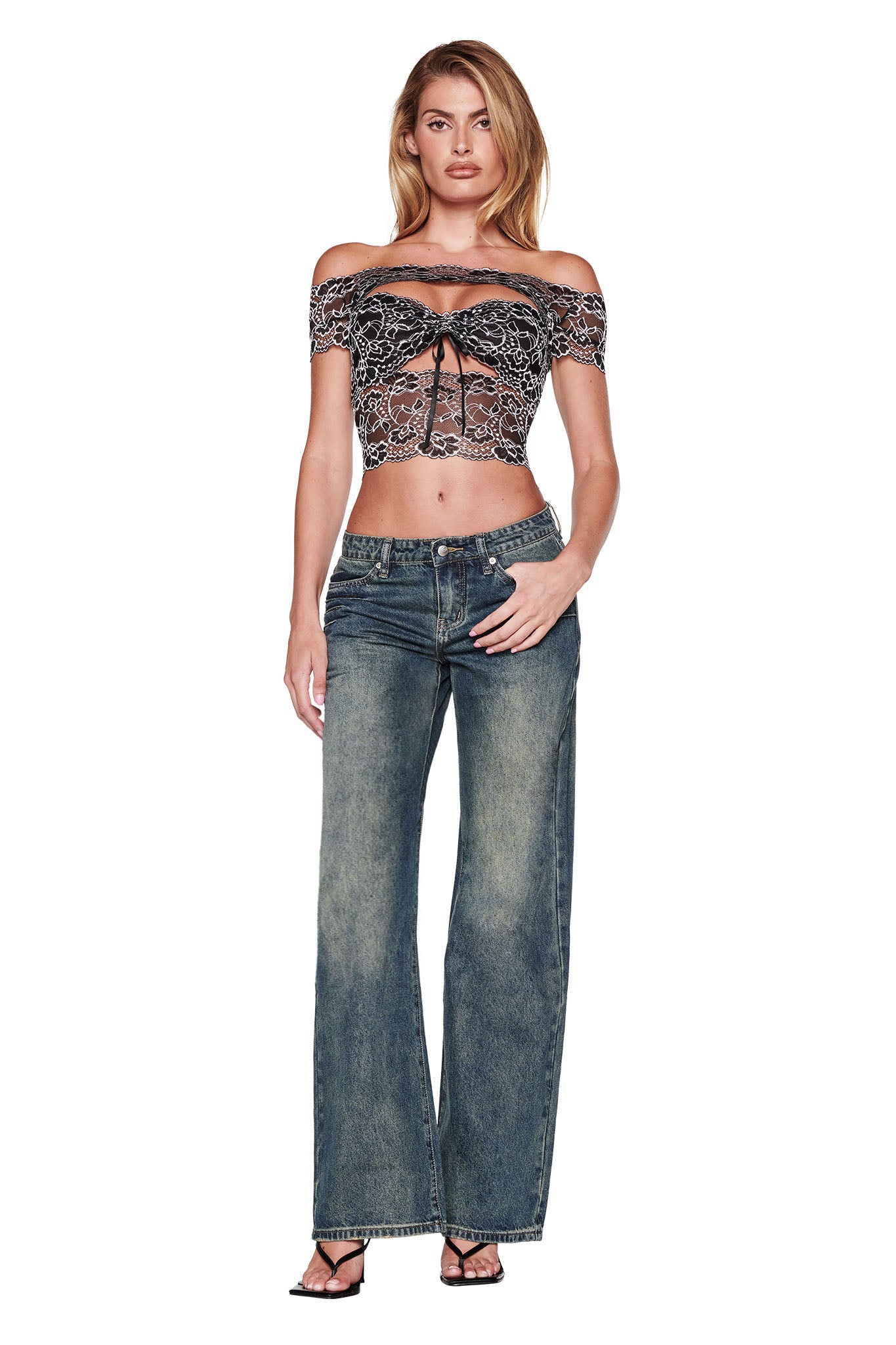 A person confidently stands with hands at their sides, wearing a black lace off-shoulder crop top and the I.AM.GIA DYLAN JEAN in blue denim. The plain white background highlights this chic ensemble.