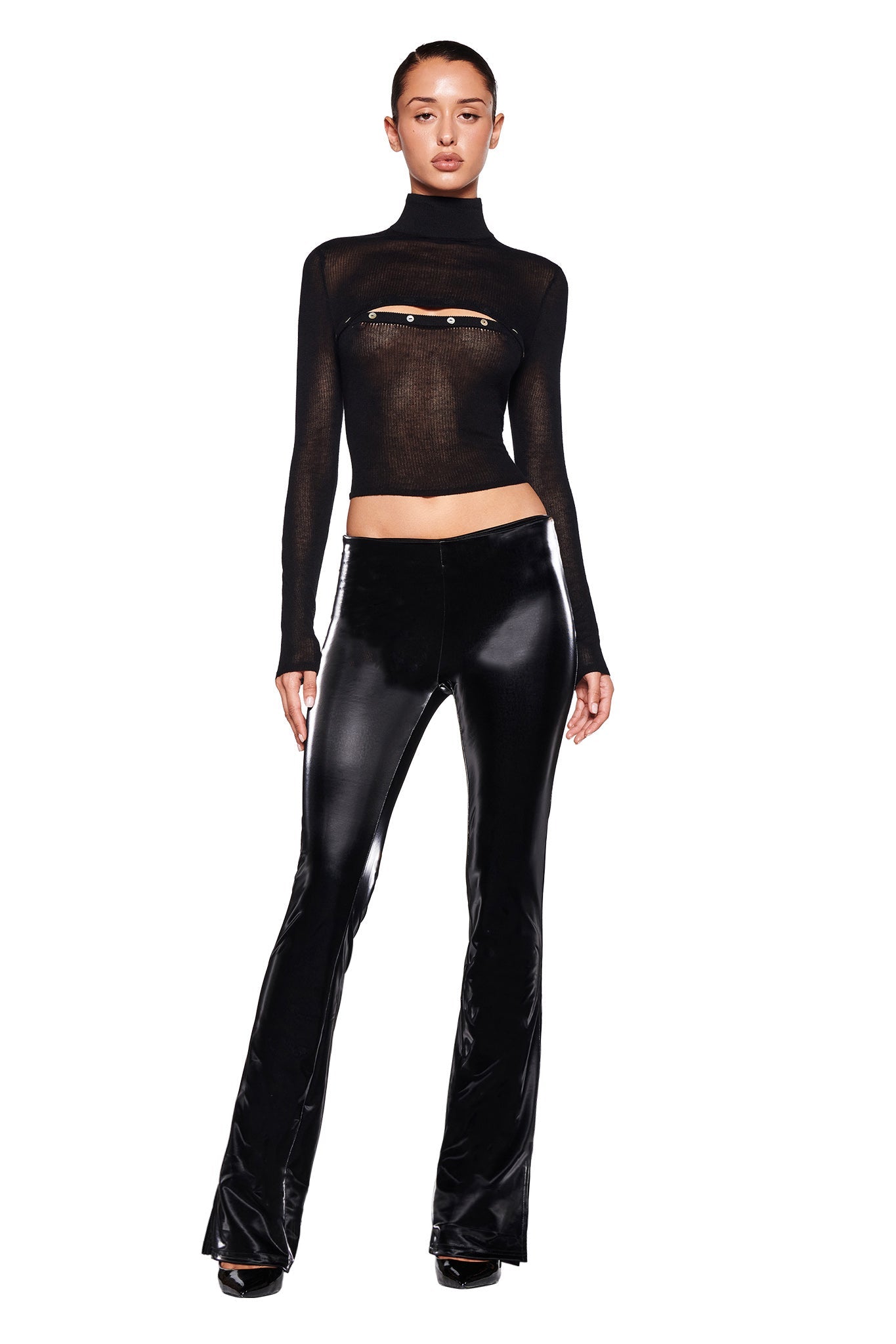 A person stands against a white background wearing a black sheer long-sleeve top with a cut-out and decorative strapping across the chest, paired with the HARLO PANT in black by I.AM.GIA and black platform shoes. The individual faces forward with a confident pose.