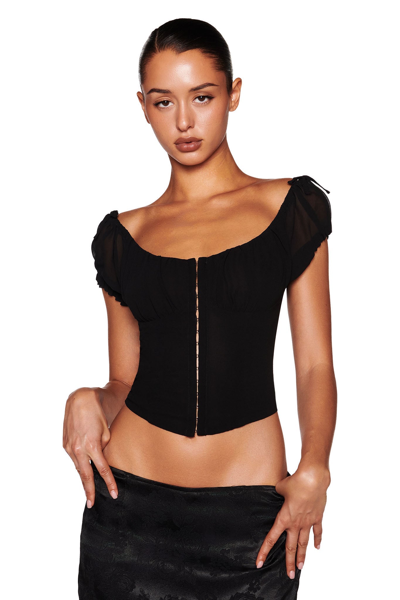 Against a white backdrop, an individual models the VALERIE GEORGETTE BUSTIER in black by I.AM.GIA. The bustier's bodycon fit accentuates their silhouette, featuring cap sleeves and a deep neckline. Their long hair is pulled back as they pose with one hand on their hip.
