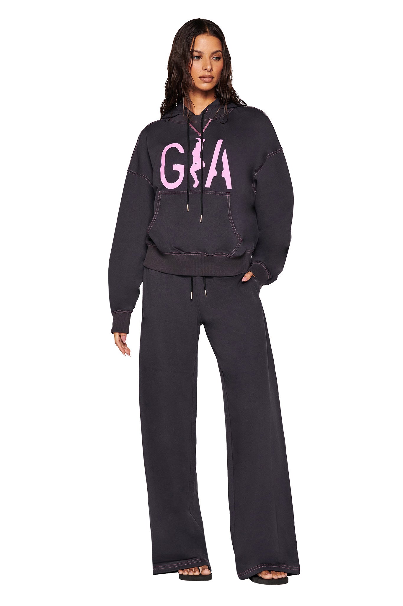 A woman wearing an I.AM.GIA black hoodie featuring a large custom "G" print on the front with hot pink contrast stitching, paired with matching SPENCER PANT - GREY in charcoal grey and black sandals. She stands against a white background, her hands casually tucked in her pockets.