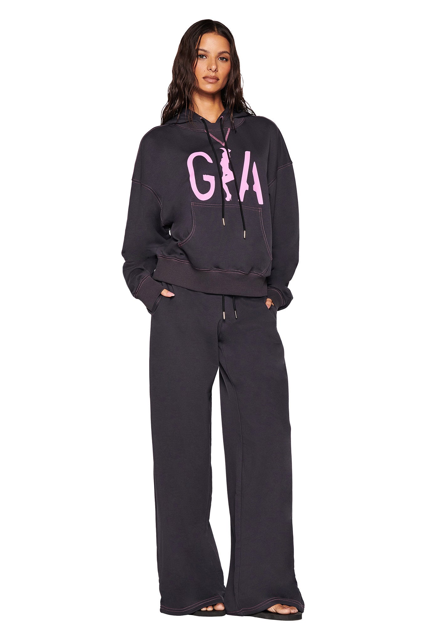 A person dressed in a dark I.AM.GIA hoodie sporting a custom pink print on the front, complemented by the SPENCER PANT in charcoal grey, which features hot pink contrast stitching. The ensemble exudes a sporty, casual vibe.