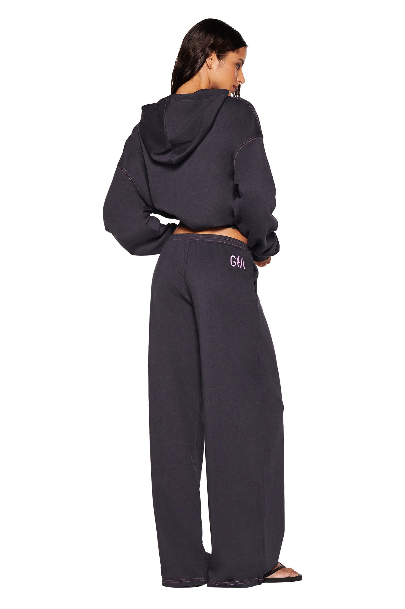 A person is dressed in the SPENCER PANT - GREY by I.AM.GIA, featuring a dark hooded tracksuit with wide-legged charcoal grey track pants. The attire is detailed with hot pink contrast stitching and has an embroidered logo on the back of the pants. They stand confidently against a white background, facing away with one hand on their hip.