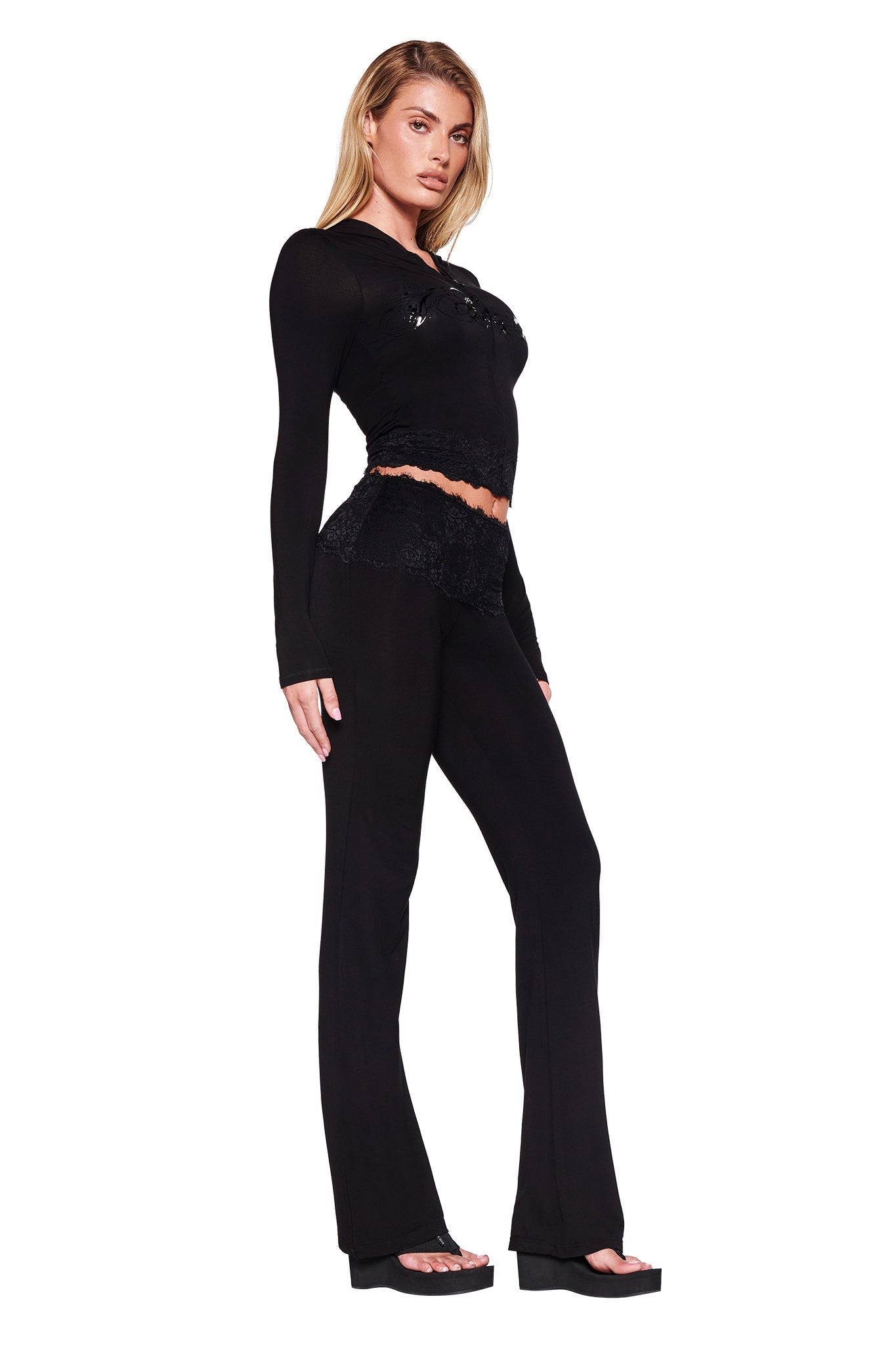 A person with long blonde hair is posing against a white background, wearing the stylish, form-fitting TARSHA JACKET - BLACK : TRUE BLACK by I.AM.GIA. The jacket features lace details at the waist and is paired with matching black pants that showcase a bodycon silhouette. They are also wearing black platform shoes.