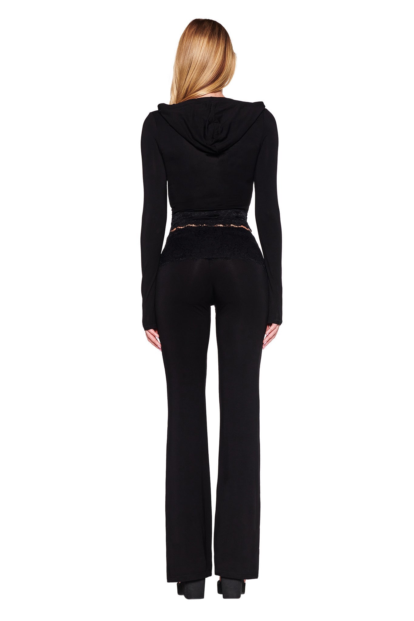 A woman with long, blonde hair is seen from the back wearing a black hooded top featuring the I.AM.GIA logo and matching black pants. She is standing against a plain white background. Her fitted TARSHA JACKET - BLACK : TRUE BLACK creates a sleek bodycon silhouette, while her pants are slightly flared at the bottom.