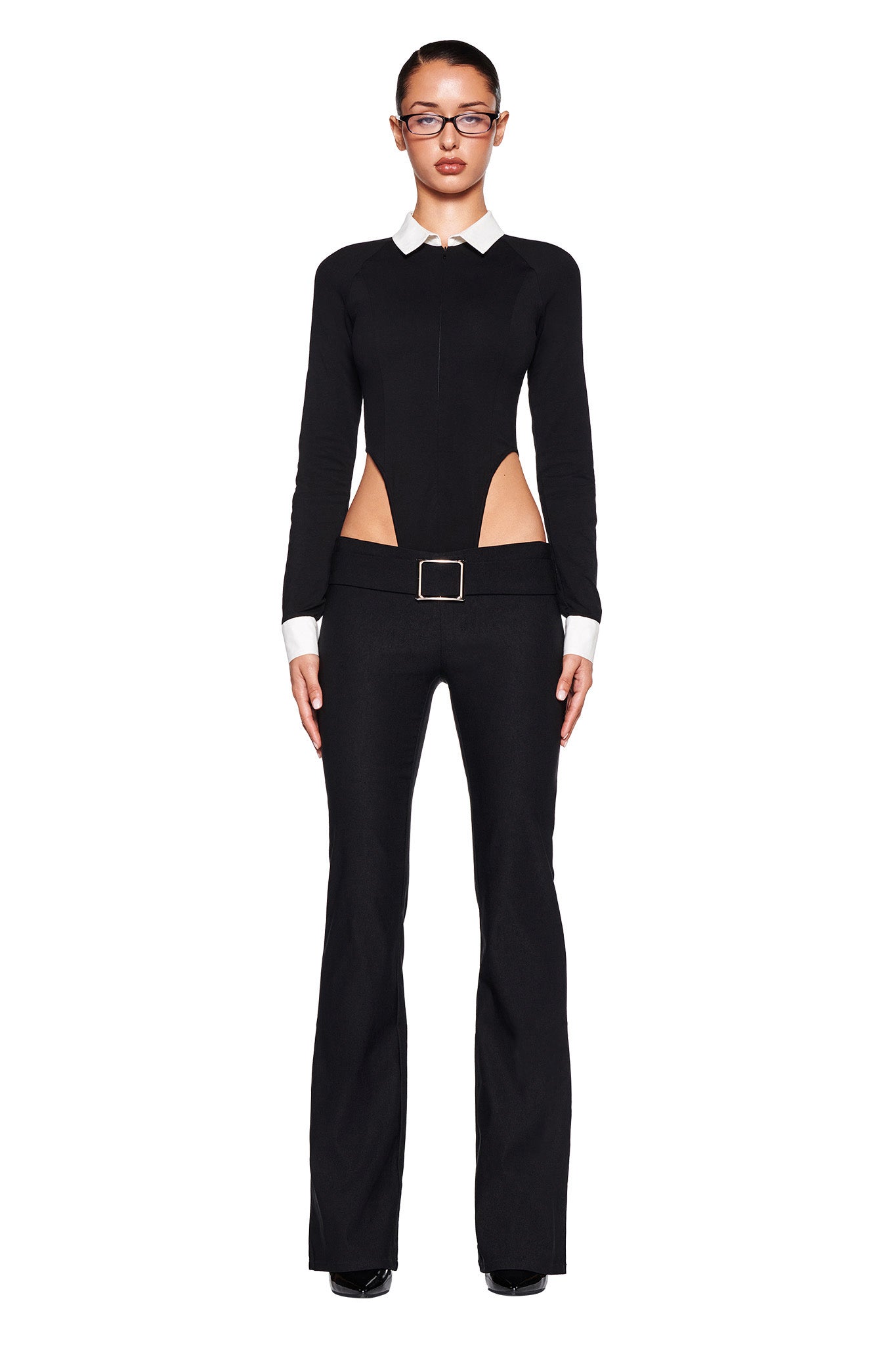 A person with a serious expression is standing and facing forward, wearing the fitted black I.AM.GIA TARA TUXEDO BODYSUIT, which features white collar cuffs, side cutouts around the waist, and a rectangular belt buckle. The long sleeve bodysuit is accessorized with glasses and black shoes against a white background.