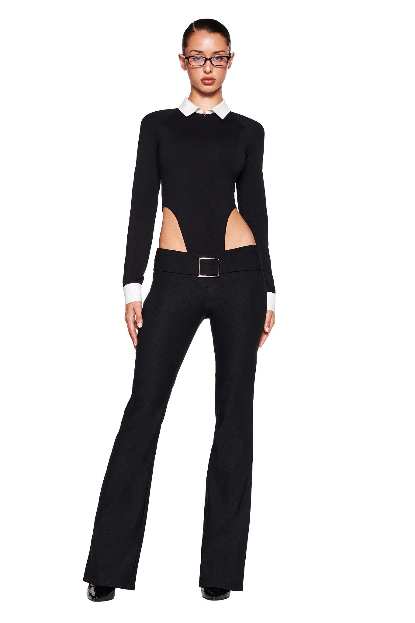 A person wearing the TARA TUXEDO BODYSUIT by I.AM.GIA, featuring black fabric, white cuffs, and a collar. The bodycon silhouette showcases distinctive cutouts at the waist and a prominent buckle on the belt. The person is also wearing glasses and black shoes, standing against a plain white background.