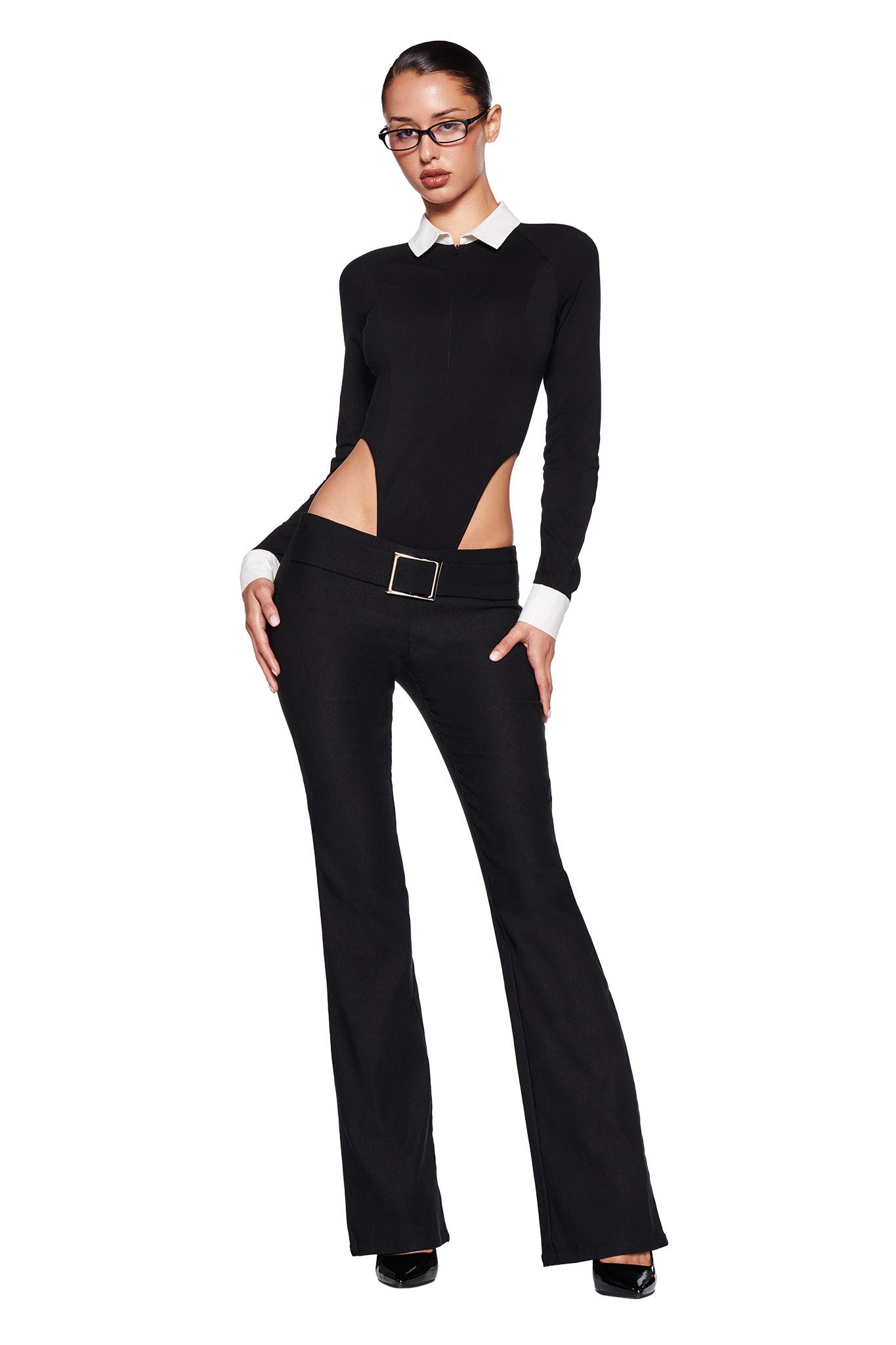 A person stands against a white background. They are wearing the I.AM.GIA TARA TUXEDO BODYSUIT, featuring a black top with cut-out sides, white collar and cuffs, paired with black belted flared pants. They have black-framed glasses and black high heels. Their hands rest on their hips.