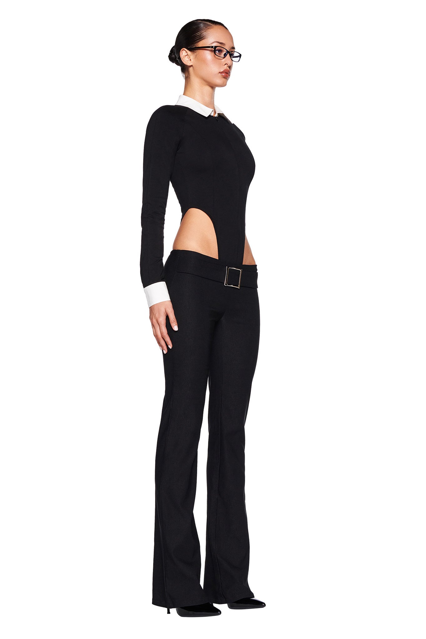A woman stands in a side profile wearing the sleek I.AM.GIA TARA TUXEDO BODYSUIT - BLACK. This long sleeve bodysuit features white cuffs, a white collar, and cut-out sides at the waist. A belt buckle accentuates her hips, while her hair is tied back and she wears glasses.
