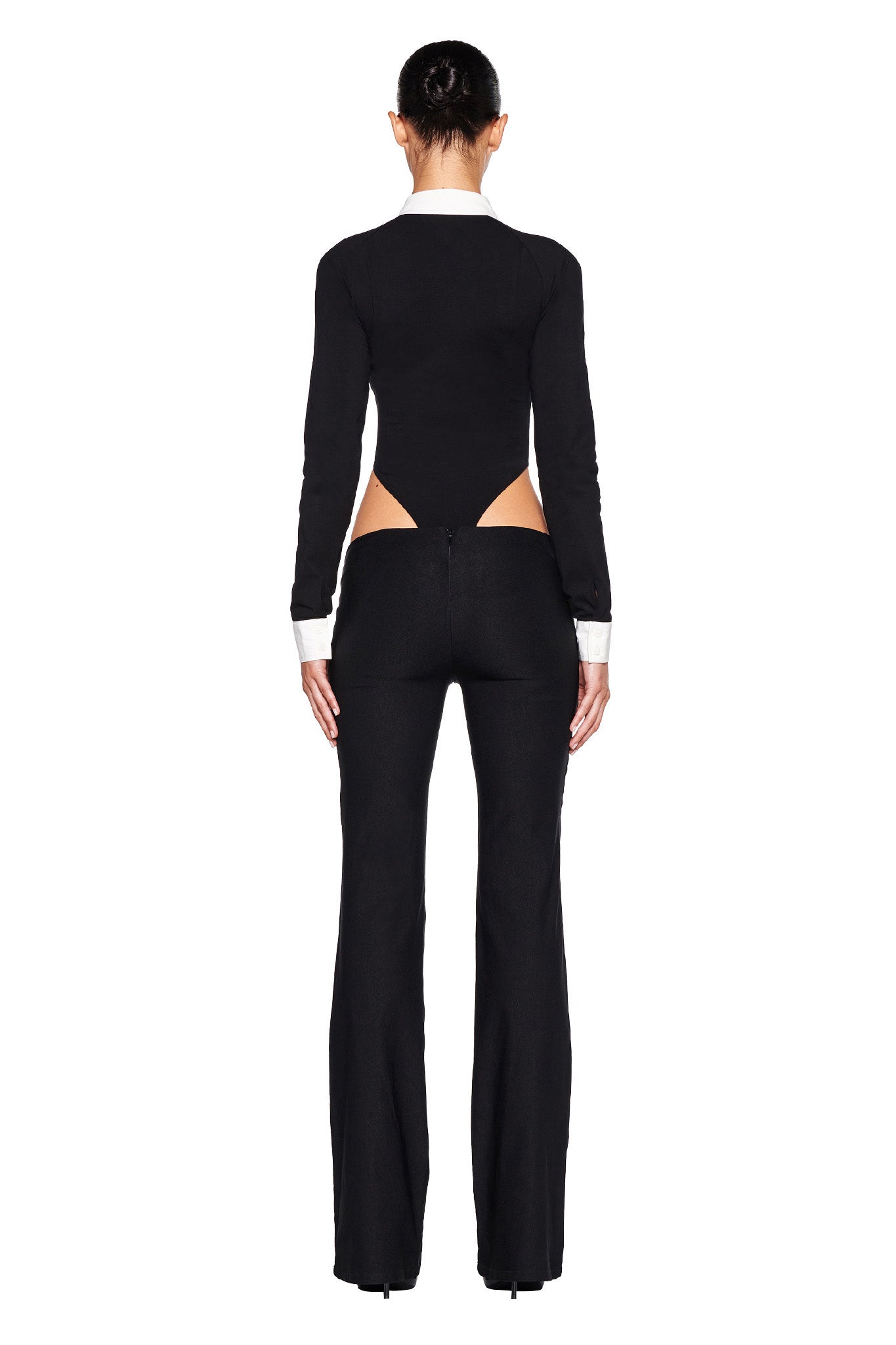 A person stands facing away, showcasing the distinctive TARA TUXEDO BODYSUIT - BLACK by I.AM.GIA, featuring a black long-sleeve top with cut-out details at the lower back and matching flared black pants. The top is accented with white cuffs and a collar, and the person's hair is tied back.