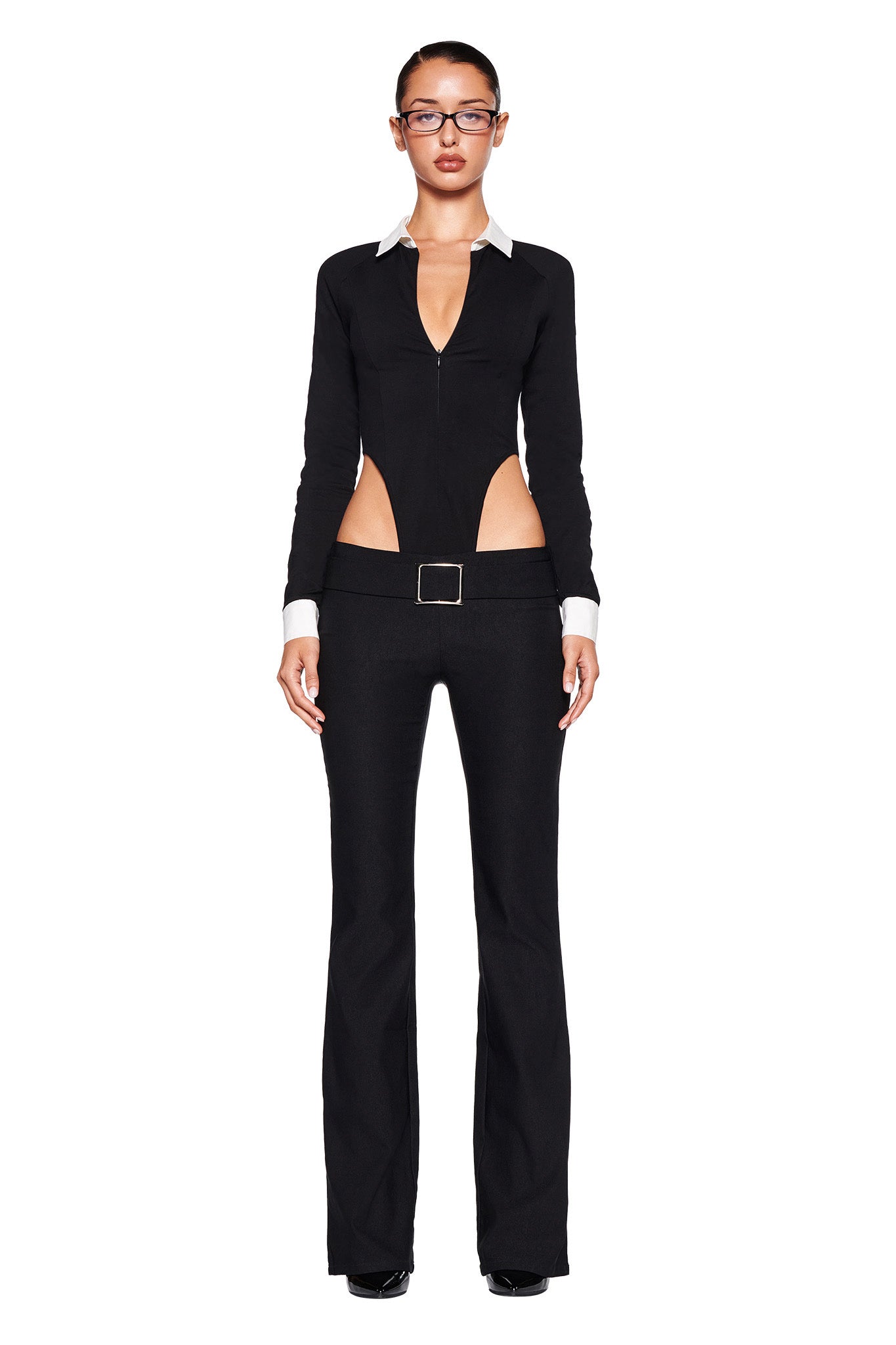 A person dressed in a stylish black outfit featuring a deep V-neck, long sleeves, and cut-out sections on the sides. They are wearing I.AM.GIA's SHARAH PANT in black, which features a low-rise waistline and flared legs, along with black shoes and glasses. The ensemble has white accents at the cuffs and collar. The background is white.